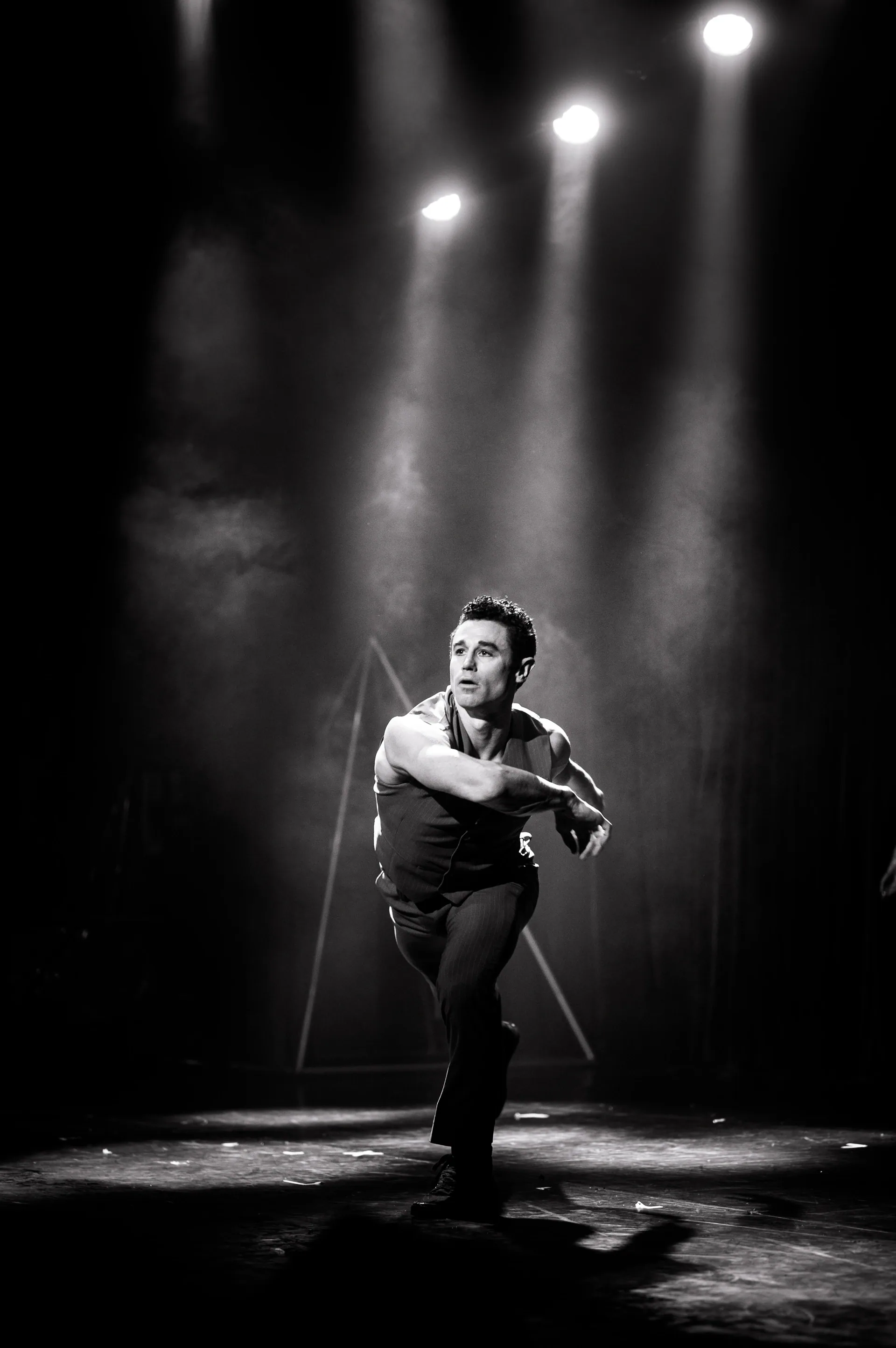 Solo performer stands under overhead spotlights with one arm extended across the body.