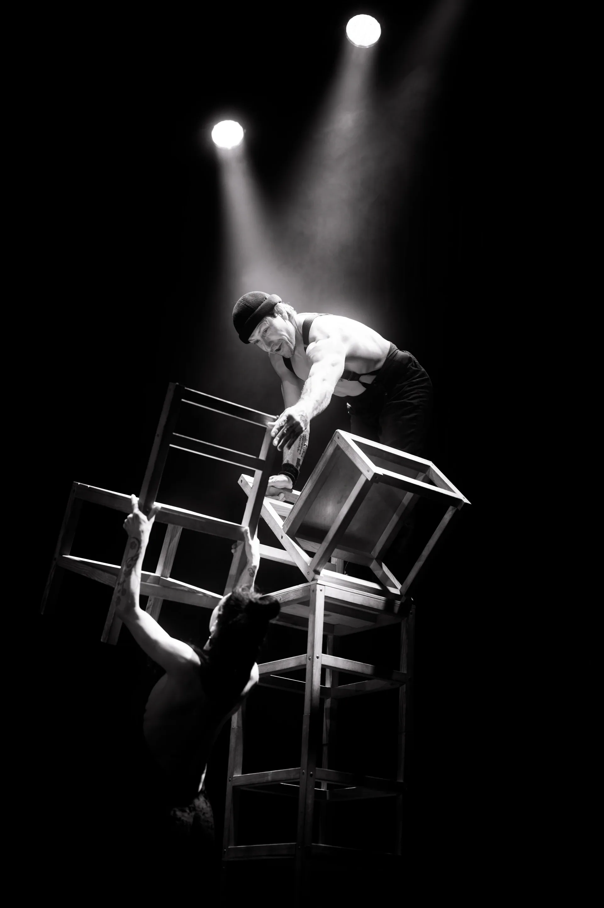 Performer balances on a stack of chairs while another steadies the structure below.