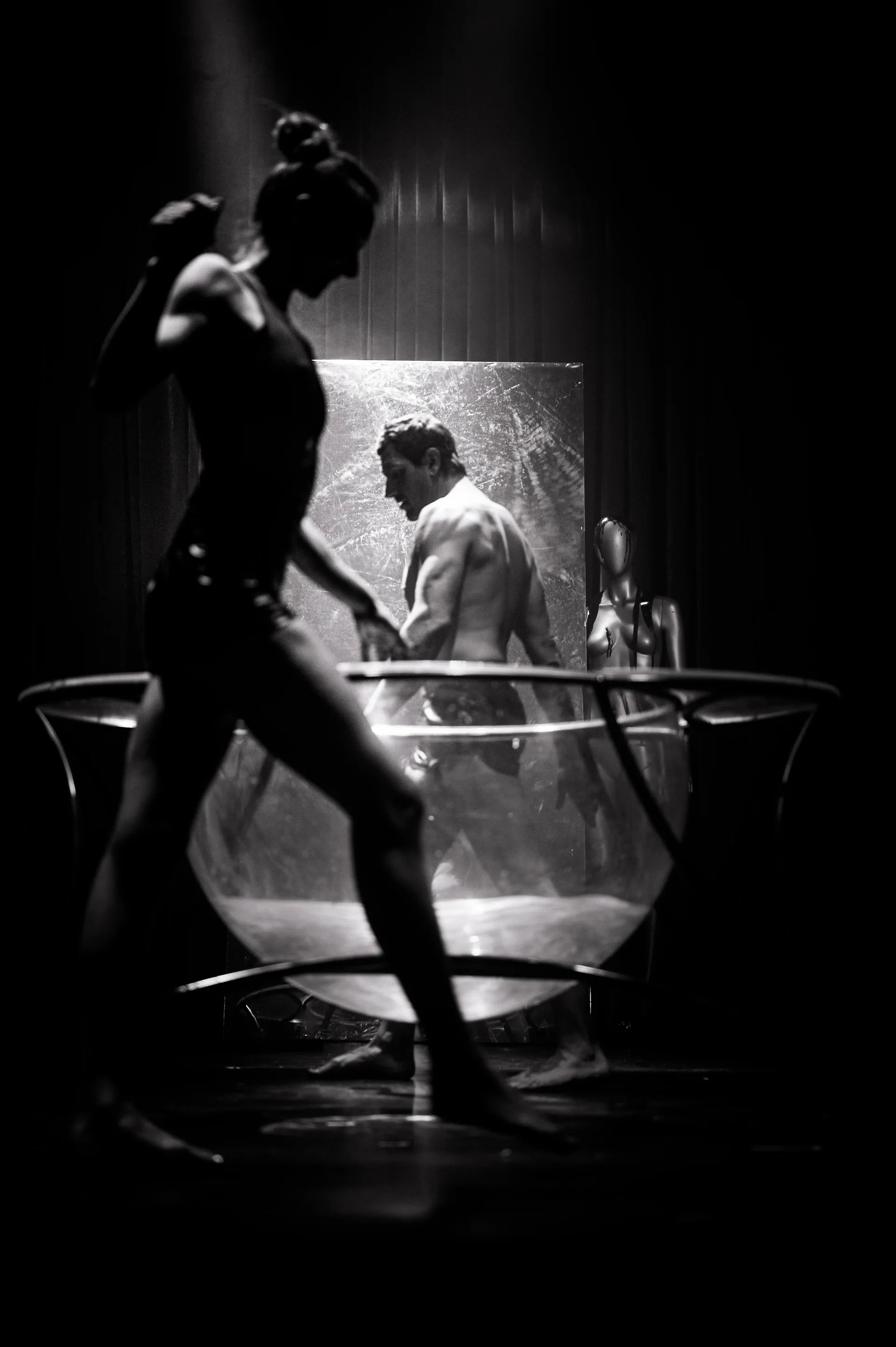 Silhouetted performer walks past a transparent water tank containing a bare-chested performer.