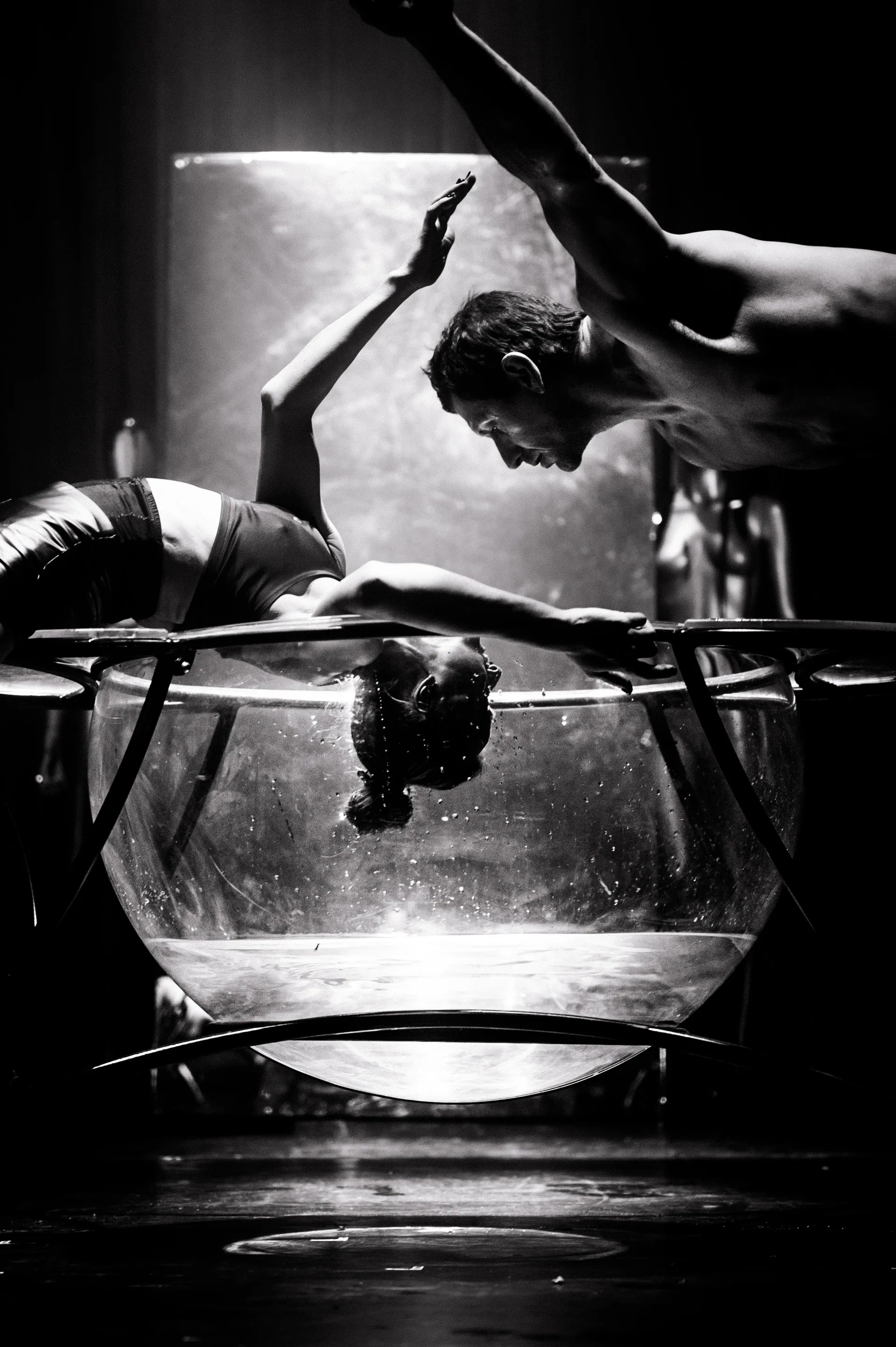 Performer bends backward over the rim of a transparent tank while another figure is visible below.