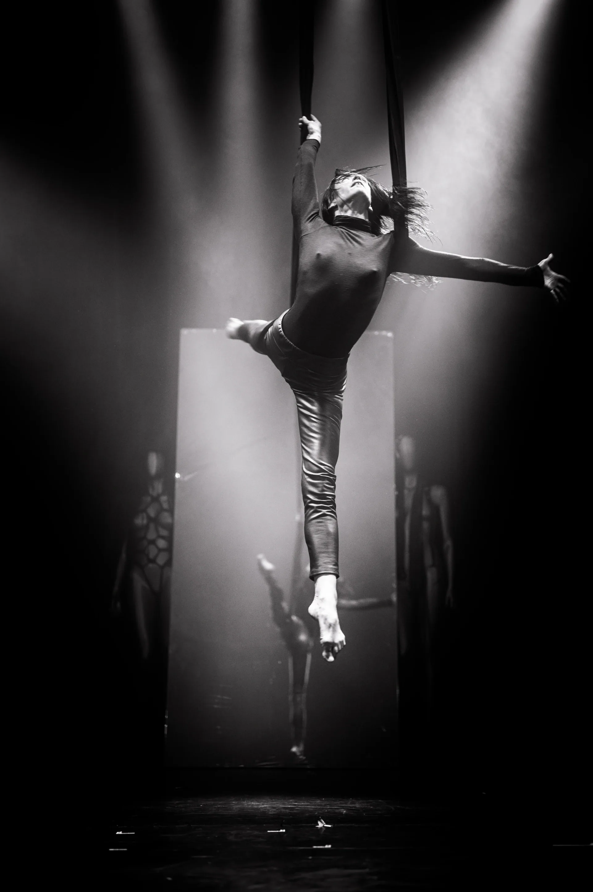 Performer extends one leg while suspended upright from an aerial apparatus.