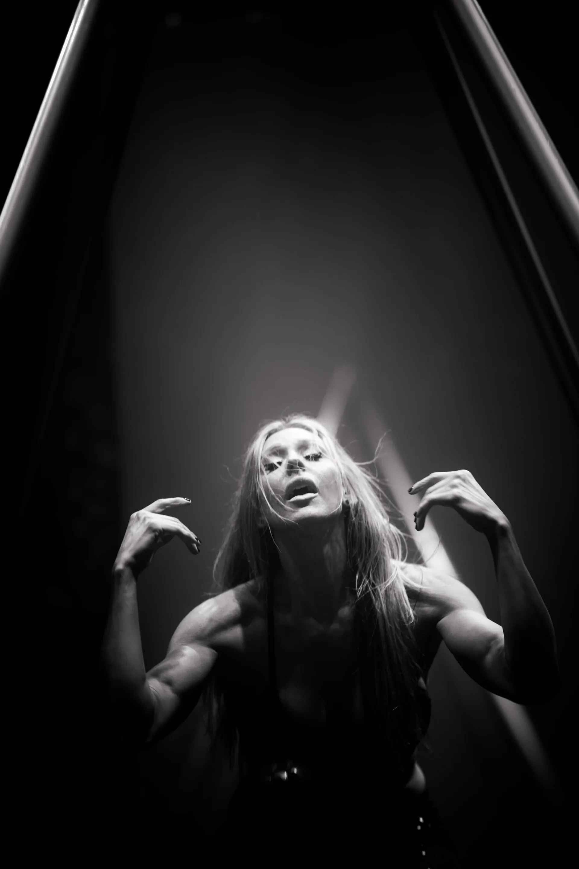 Long-haired performer leans backward with raised hands under stage light.