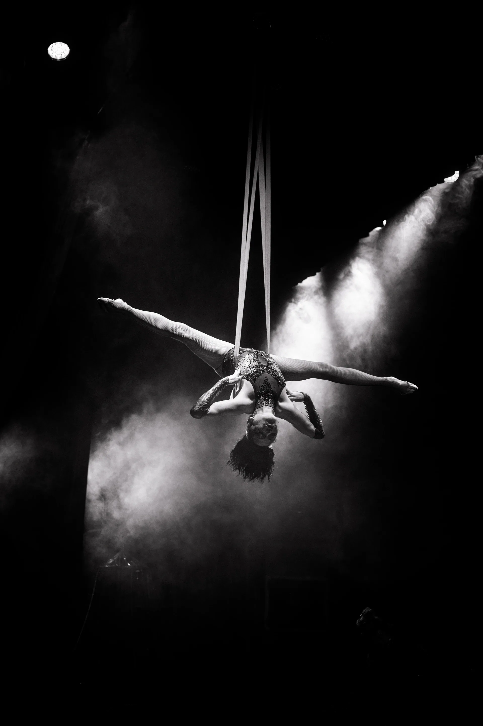 Aerial performer hangs upside down with legs extended into a split amid stage haze.