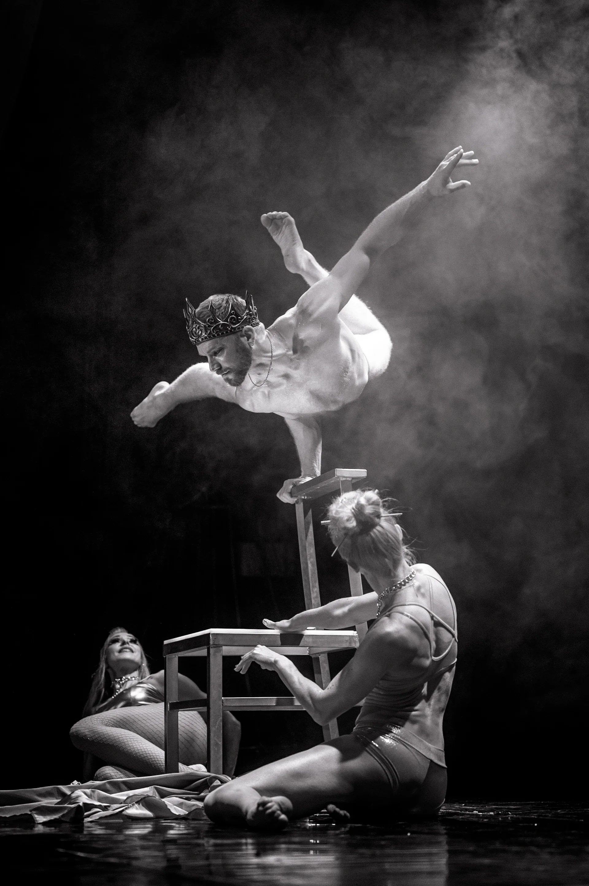 Three performers form an acrobatic tableau around a chair under a smoky spotlight.