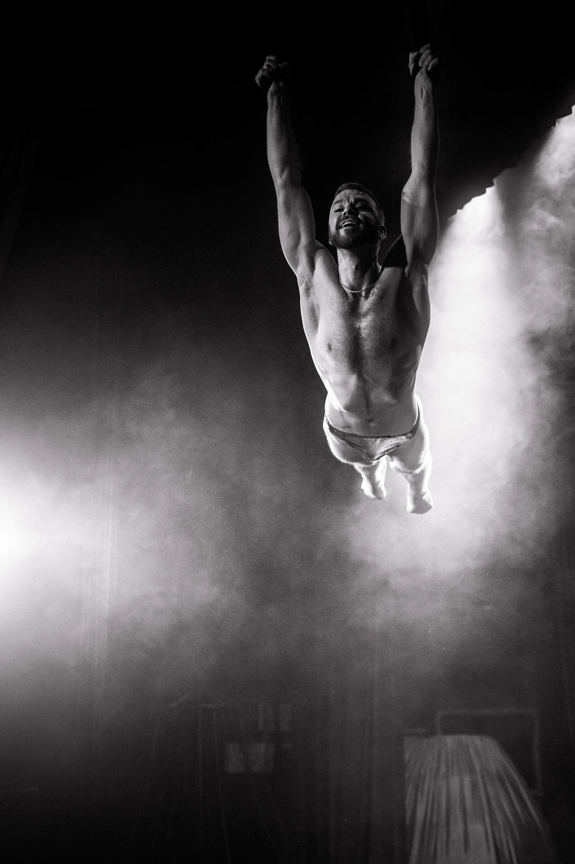 Aerial performer hangs vertically with arms extended overhead amid stage haze.