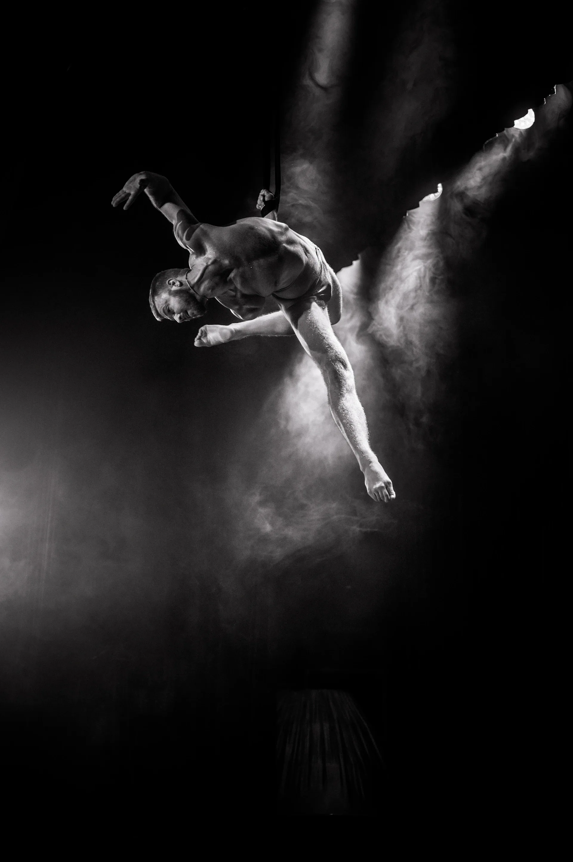 Aerial performer holds a horizontal pose in a beam of smoky light.