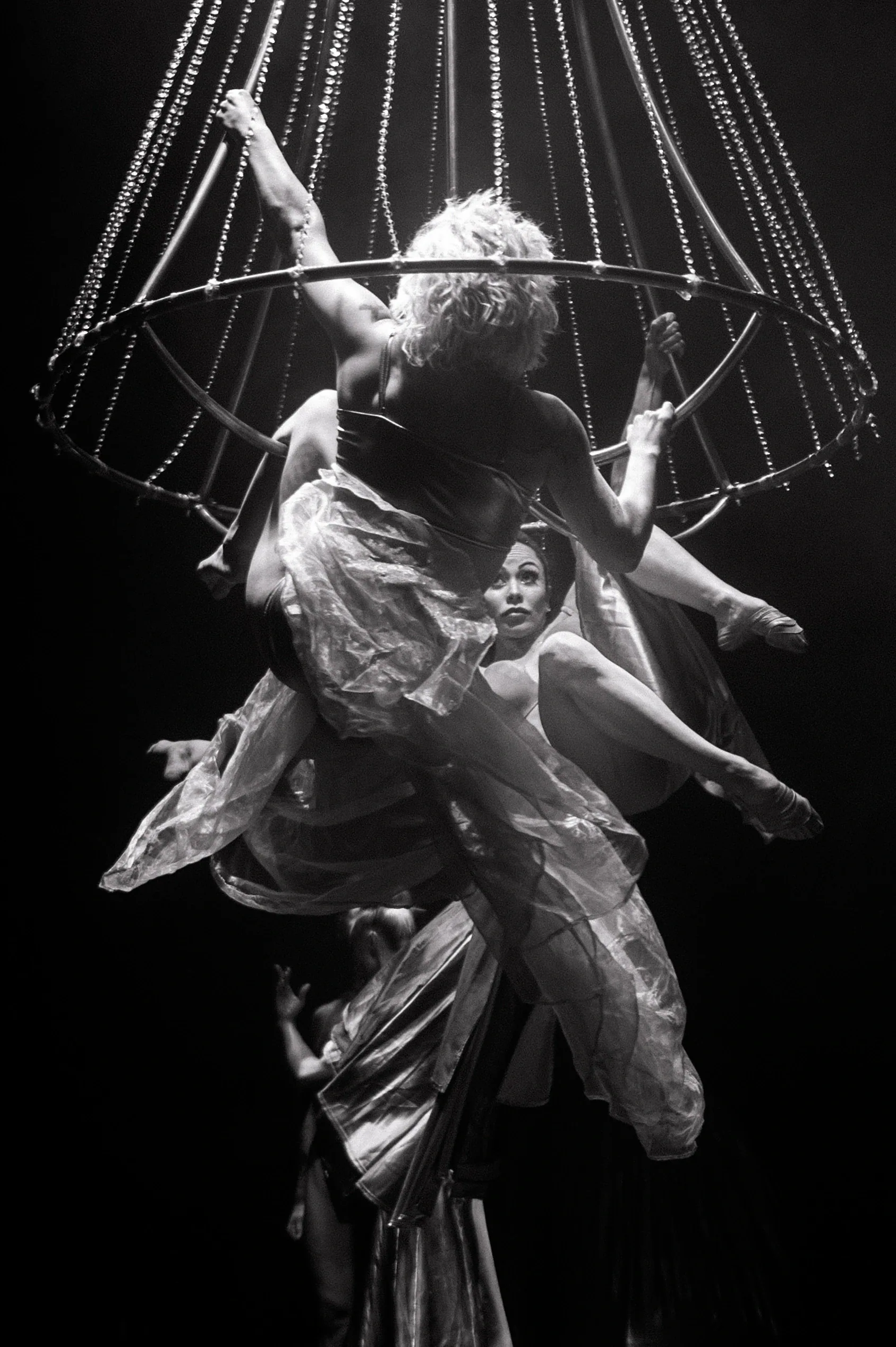Performers gather around a suspended cage-like metal frame draped with fabric.