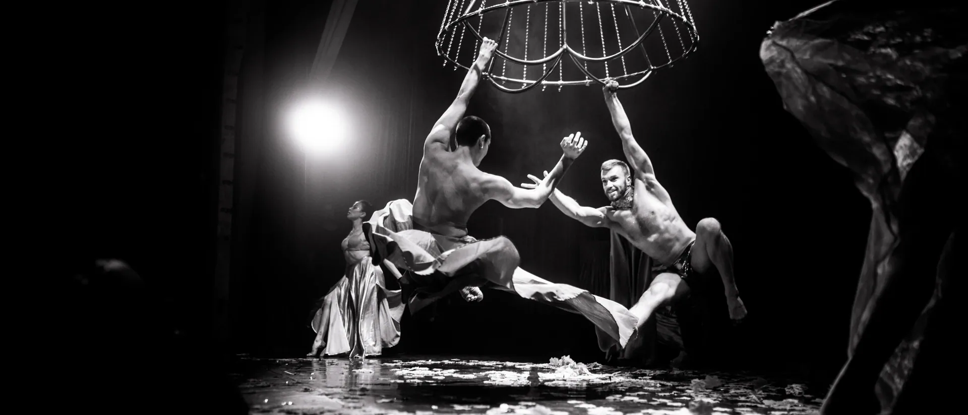 Performers reach toward one another beneath a hanging frame on a confetti-covered stage.