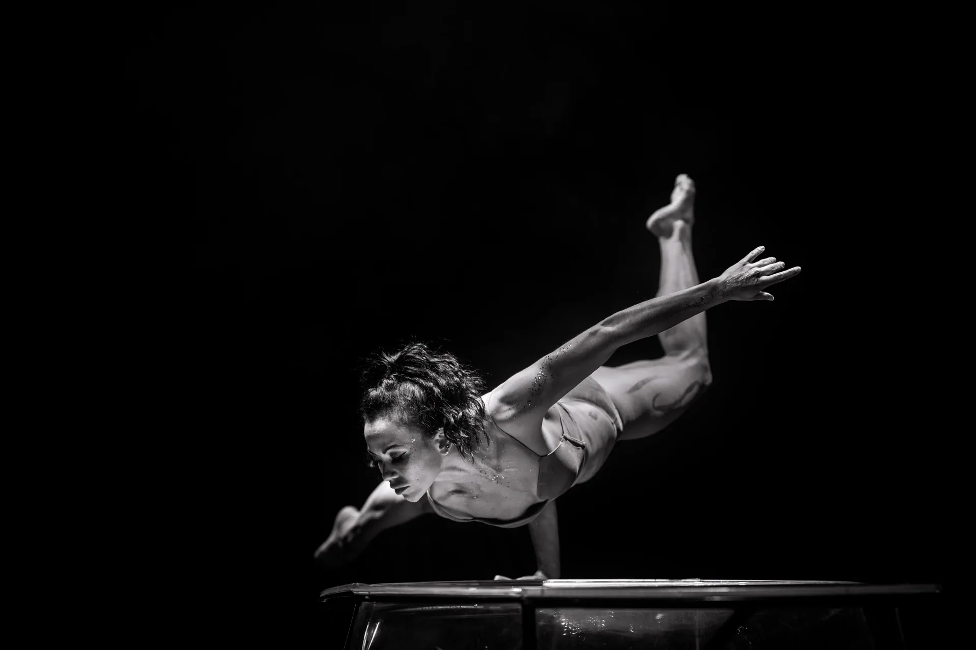 Contortionist balances on a low transparent platform with one leg extended high behind.
