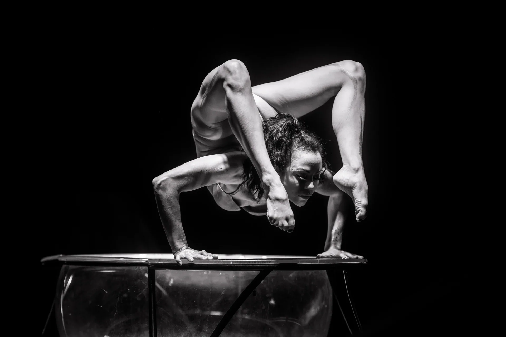 Contortionist balances on a low transparent platform with both legs curved over the shoulders.