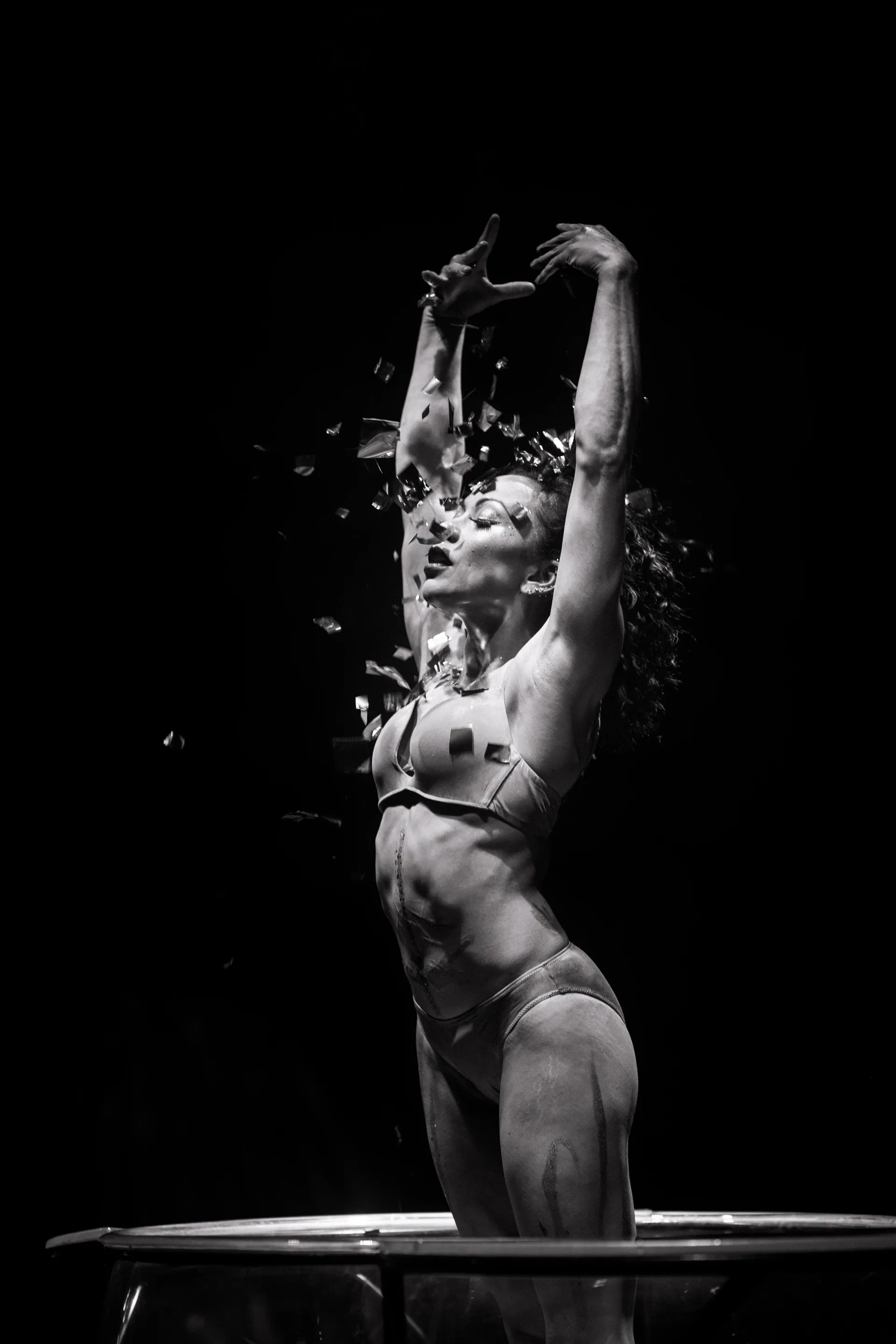 Performer stands in a transparent tank with arms raised as water splashes upward.