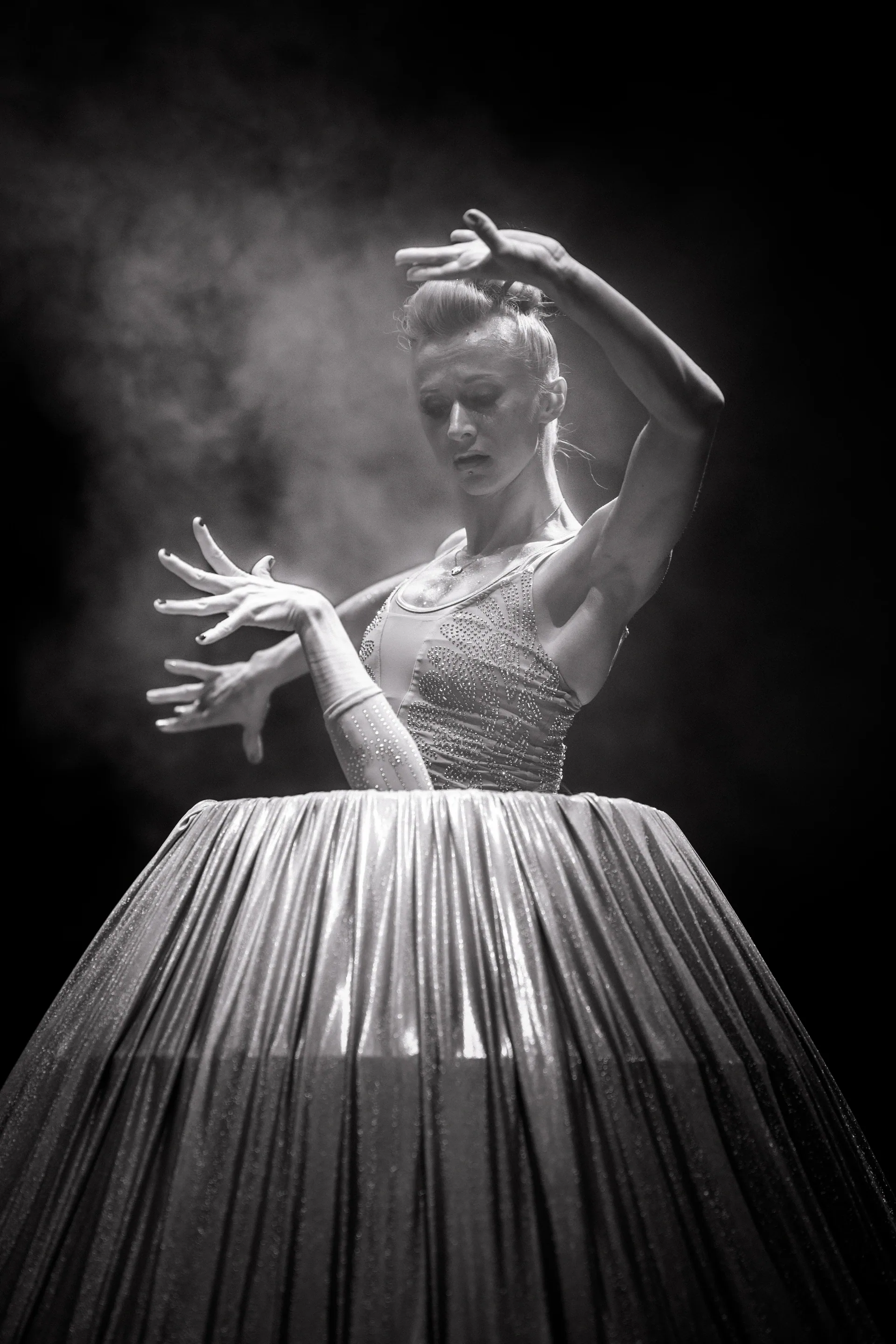 Performer gestures with both hands from behind a pleated circular fabric panel.