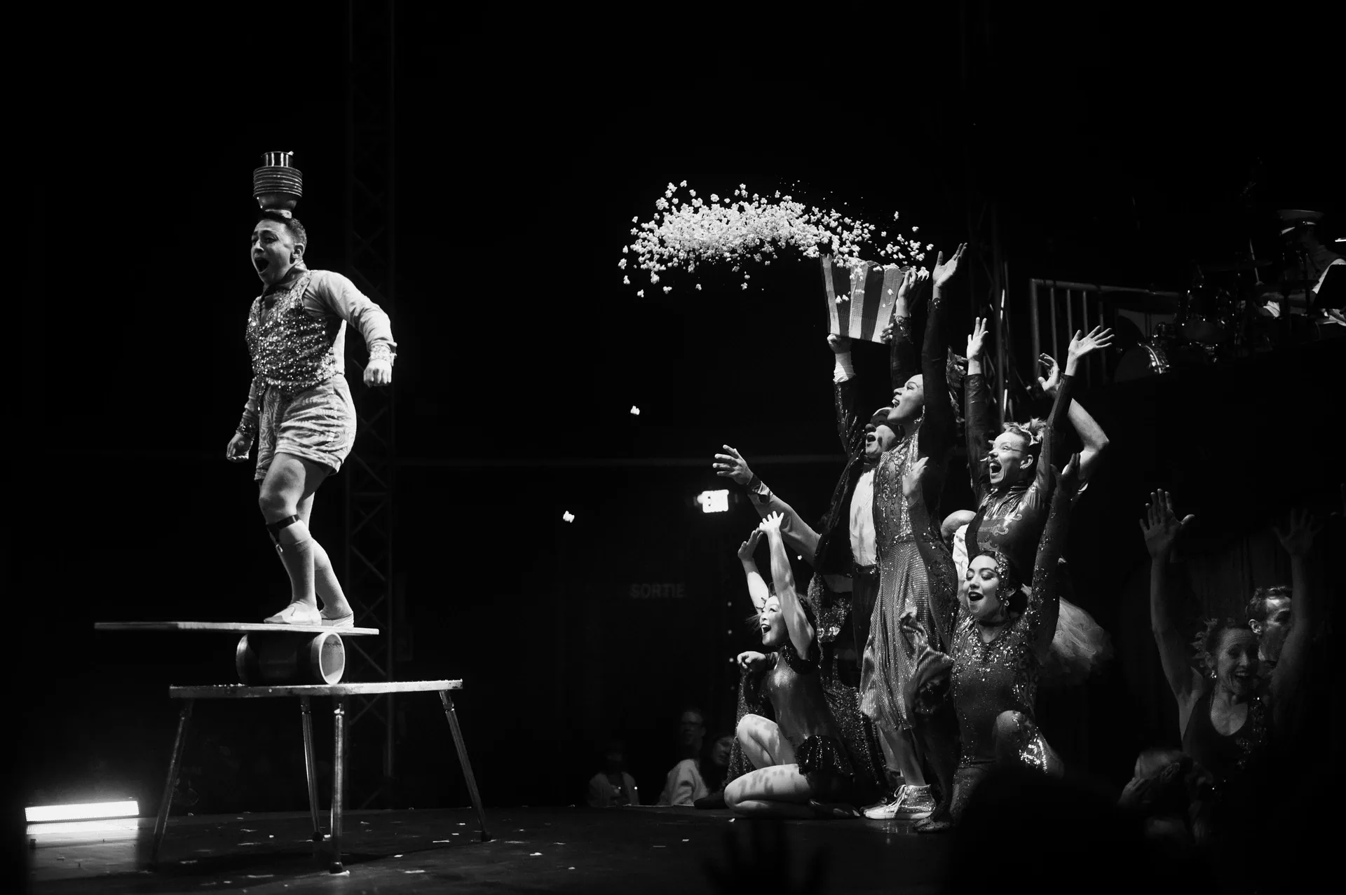 Performer balances on a small platform as an ensemble tosses white objects into the air.