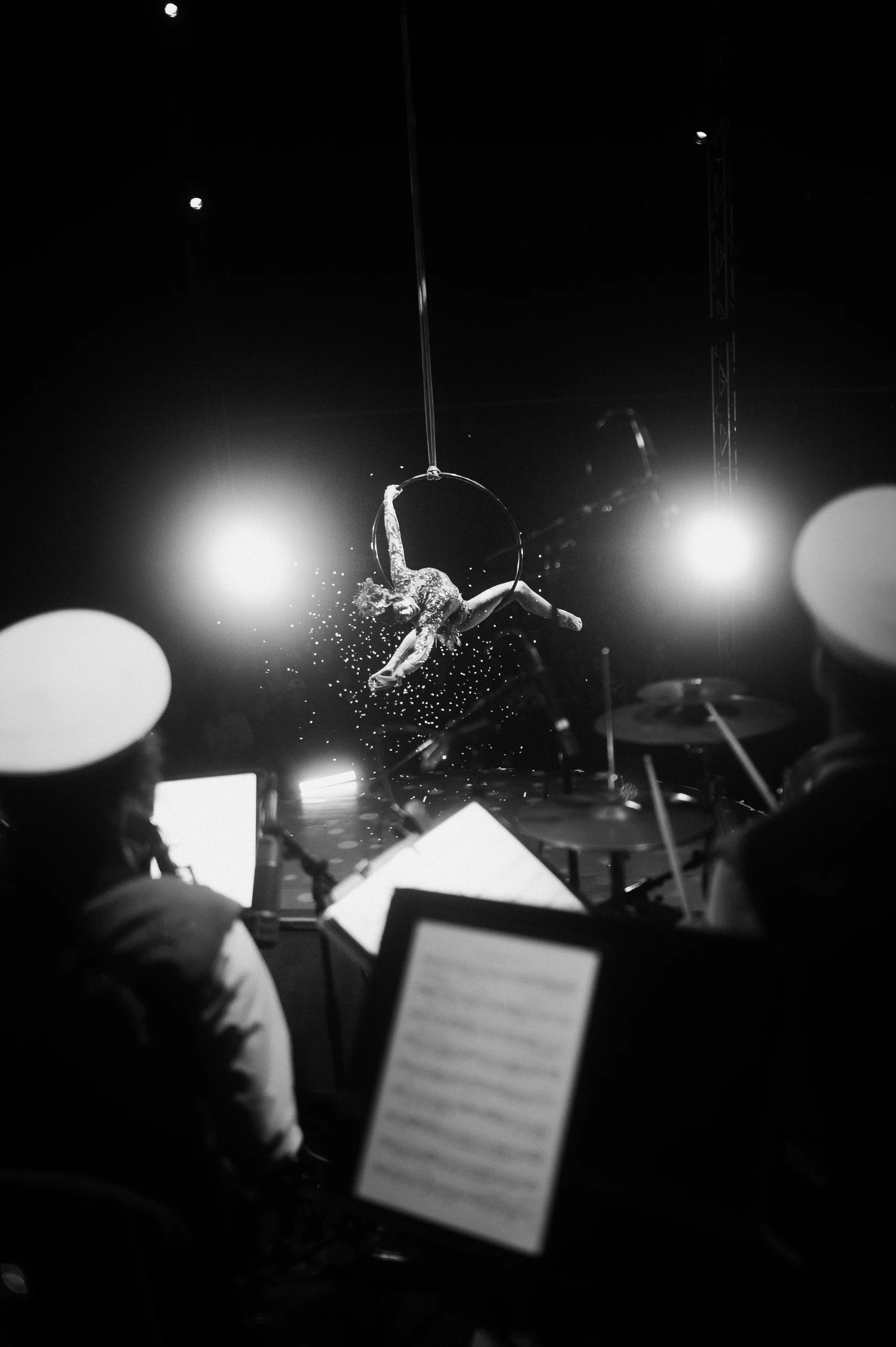 Aerialist hangs from a hoop above orchestra musicians and a music stand.