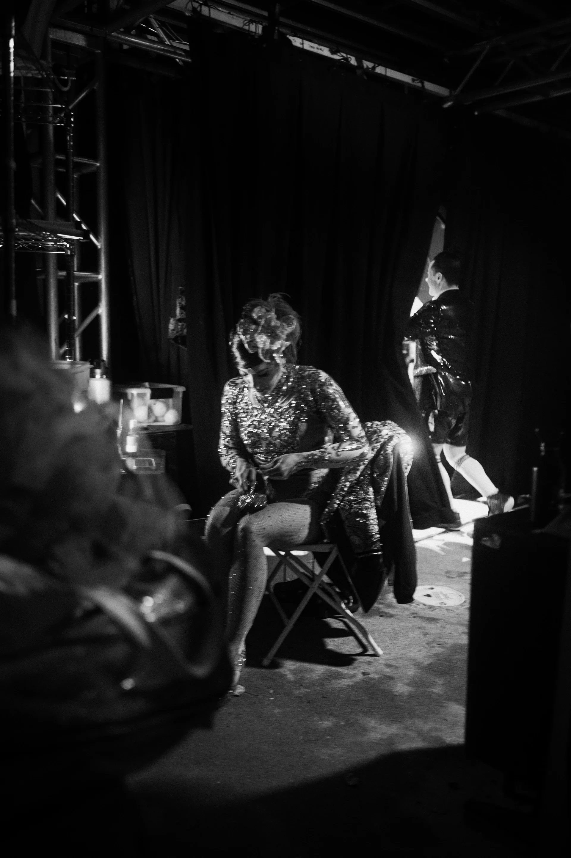 Costumed performer sits beside a stage curtain while another figure stands behind.