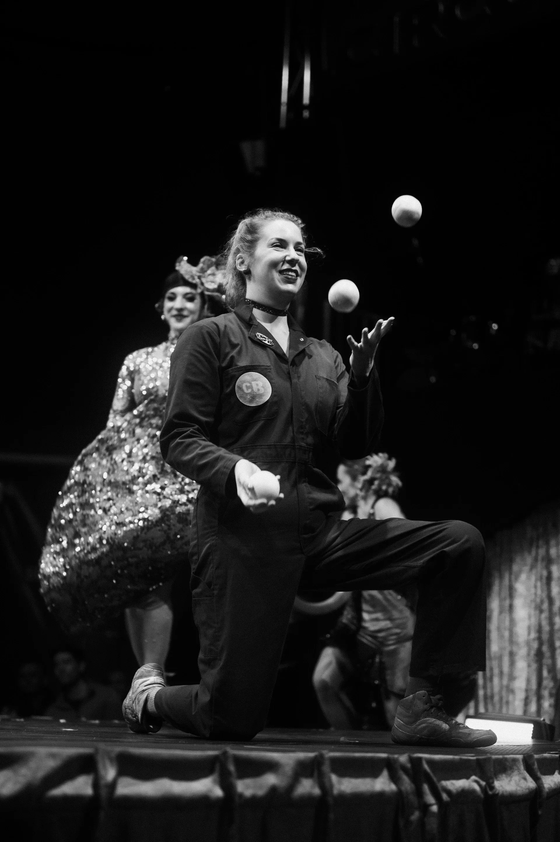 Kneeling juggler keeps several balls in the air with another performer behind.