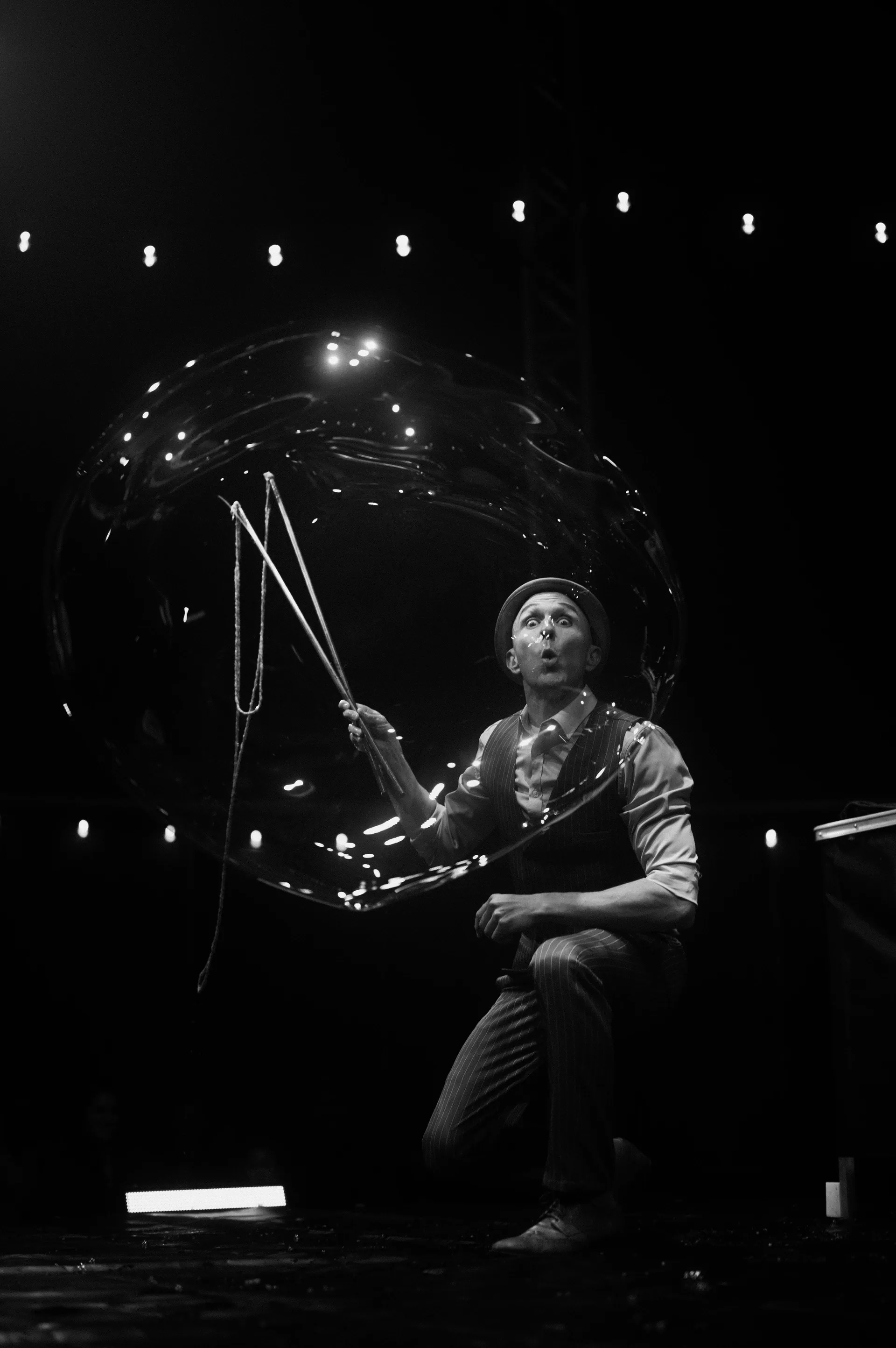 Kneeling performer manipulates a large clear sphere beneath stage lights.