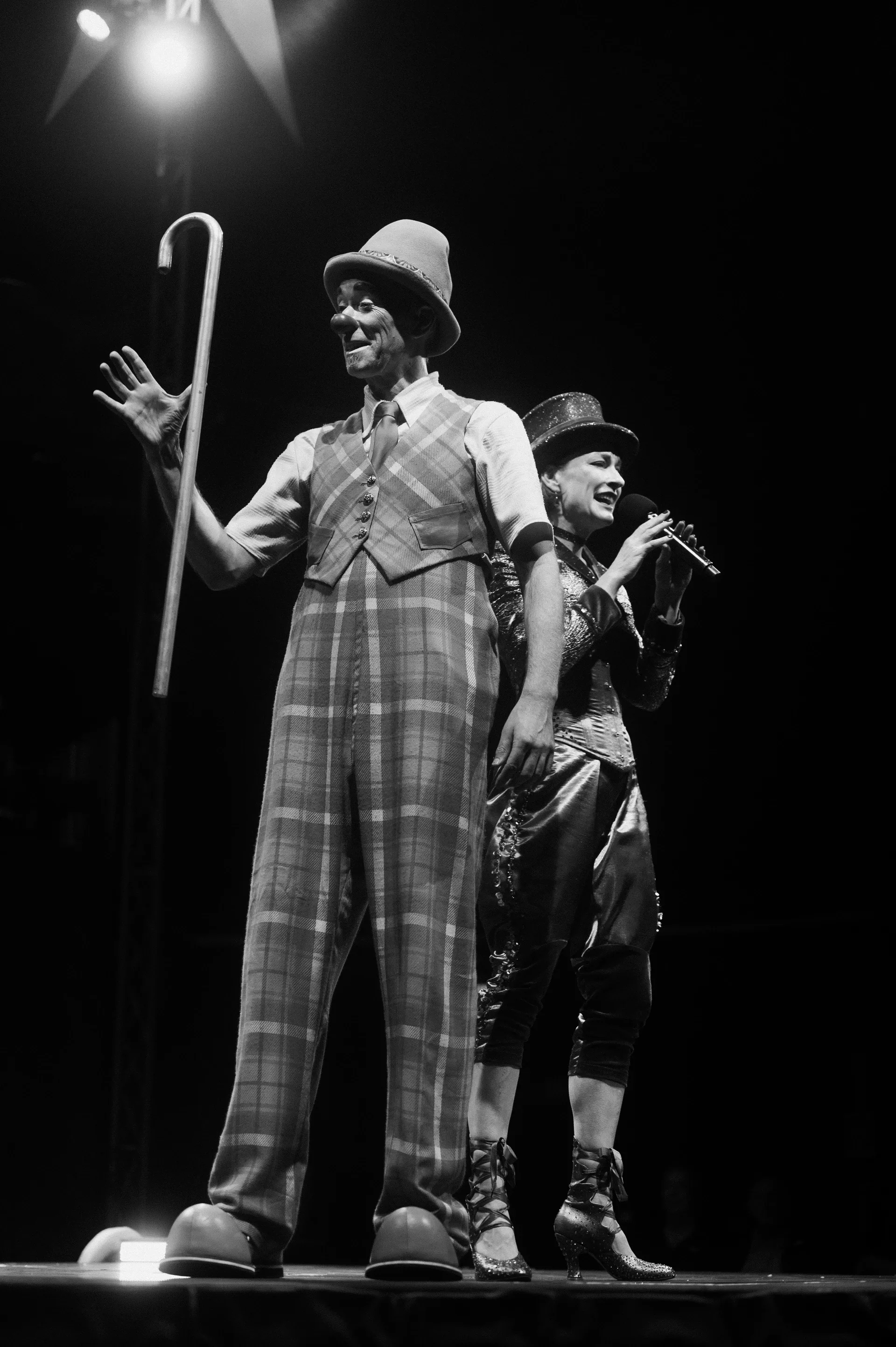Two performers in tall plaid costumes stand together, one holding a curved cane.