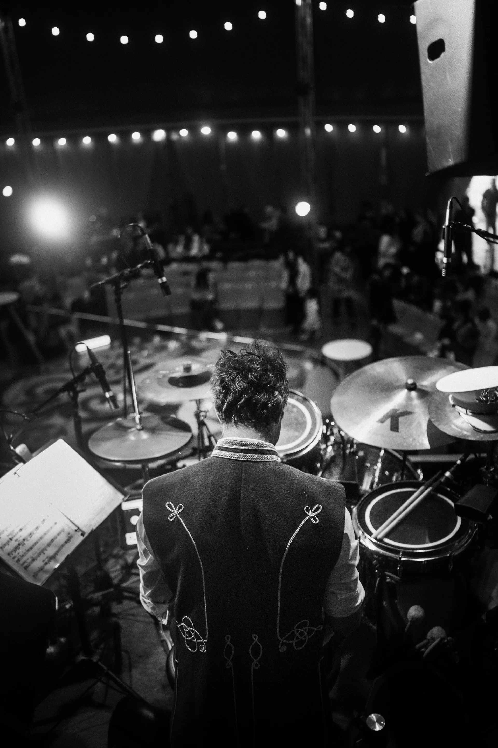 Drummer is seen from behind at a kit beneath strings of lights.