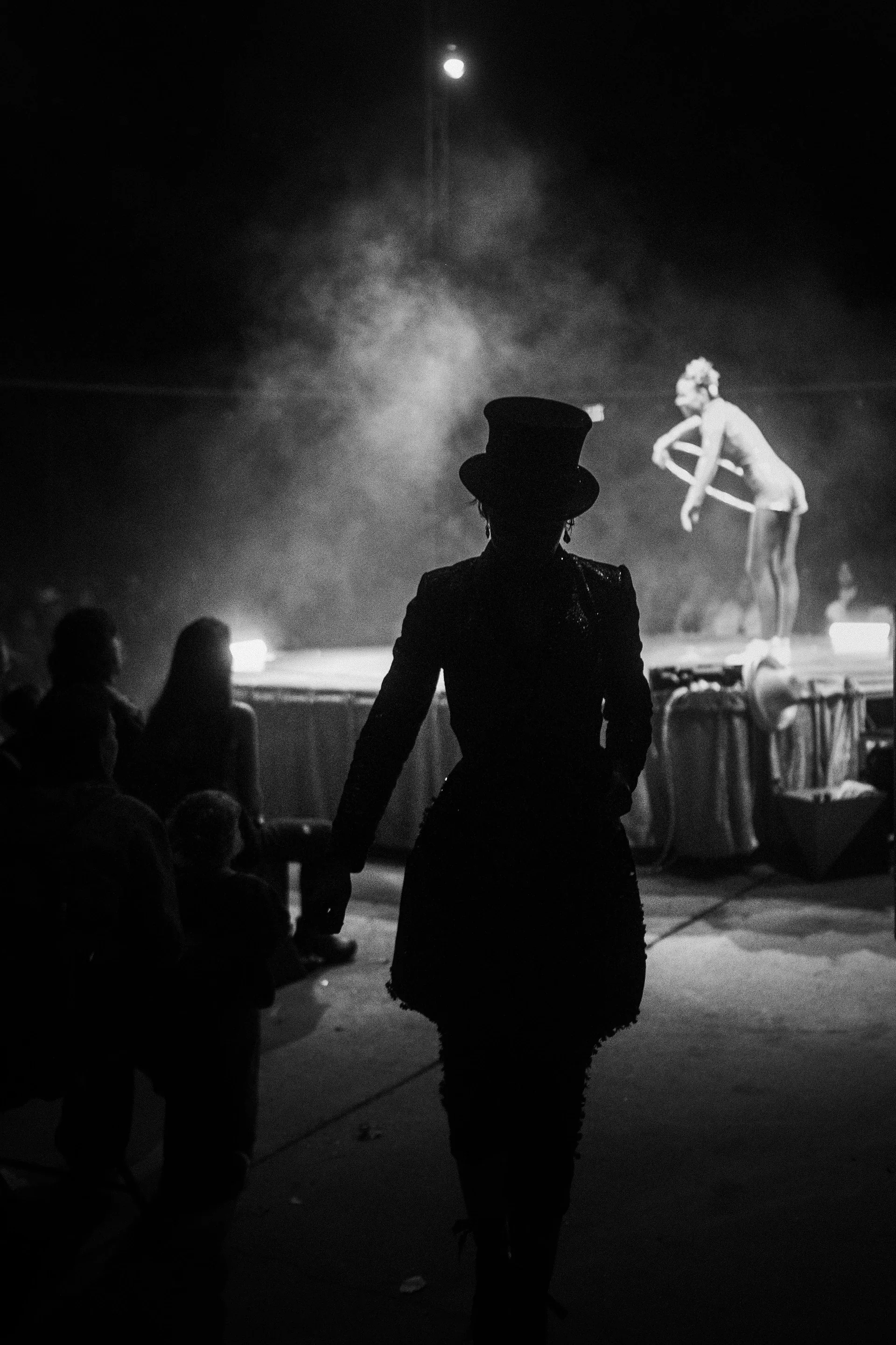 Silhouetted performer in a top hat watches an act across the stage.