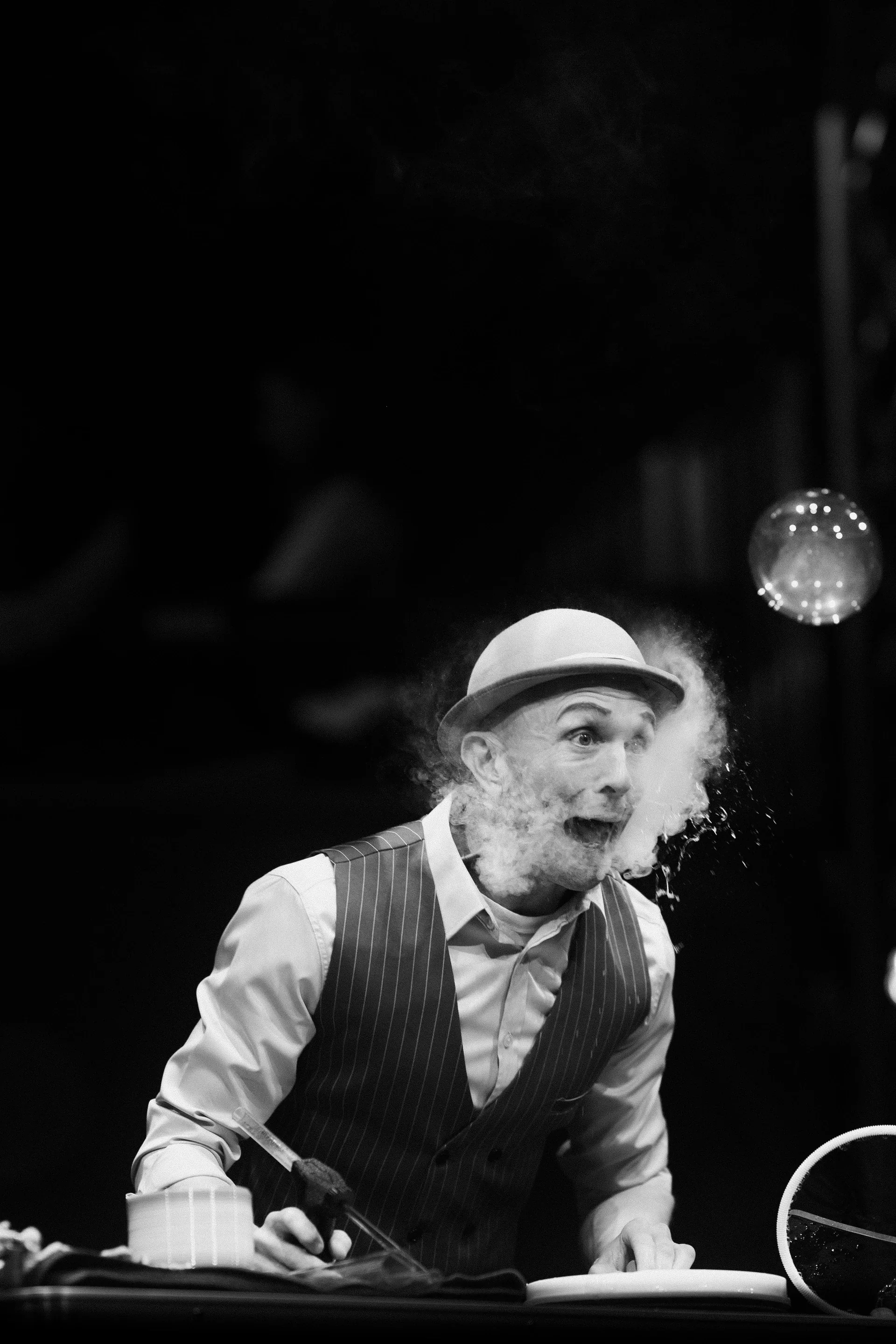 Costumed performer leans over a table while exhaling a visible cloud.