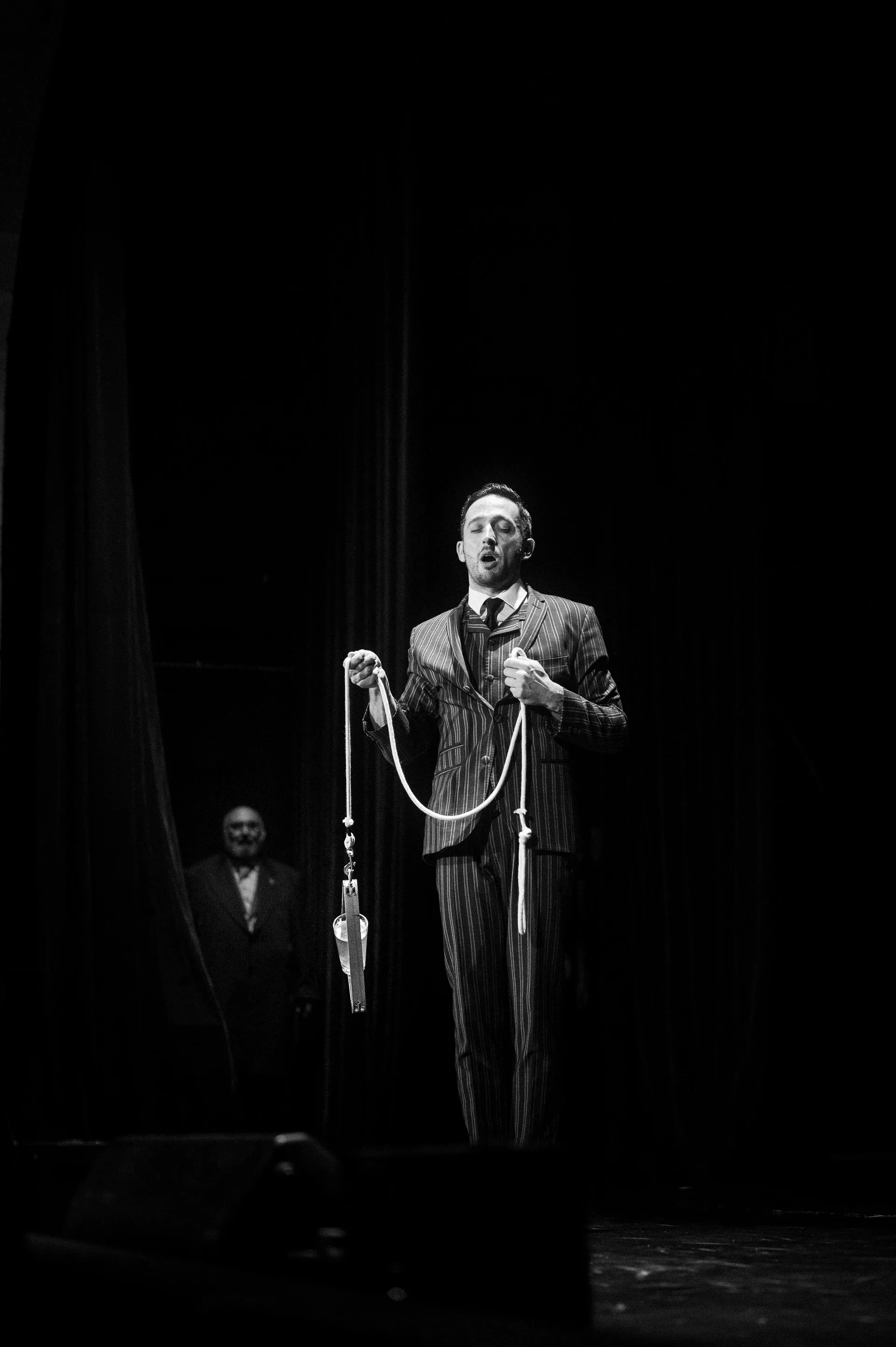 Performer in a suit holds a looped rope beneath a spotlight.