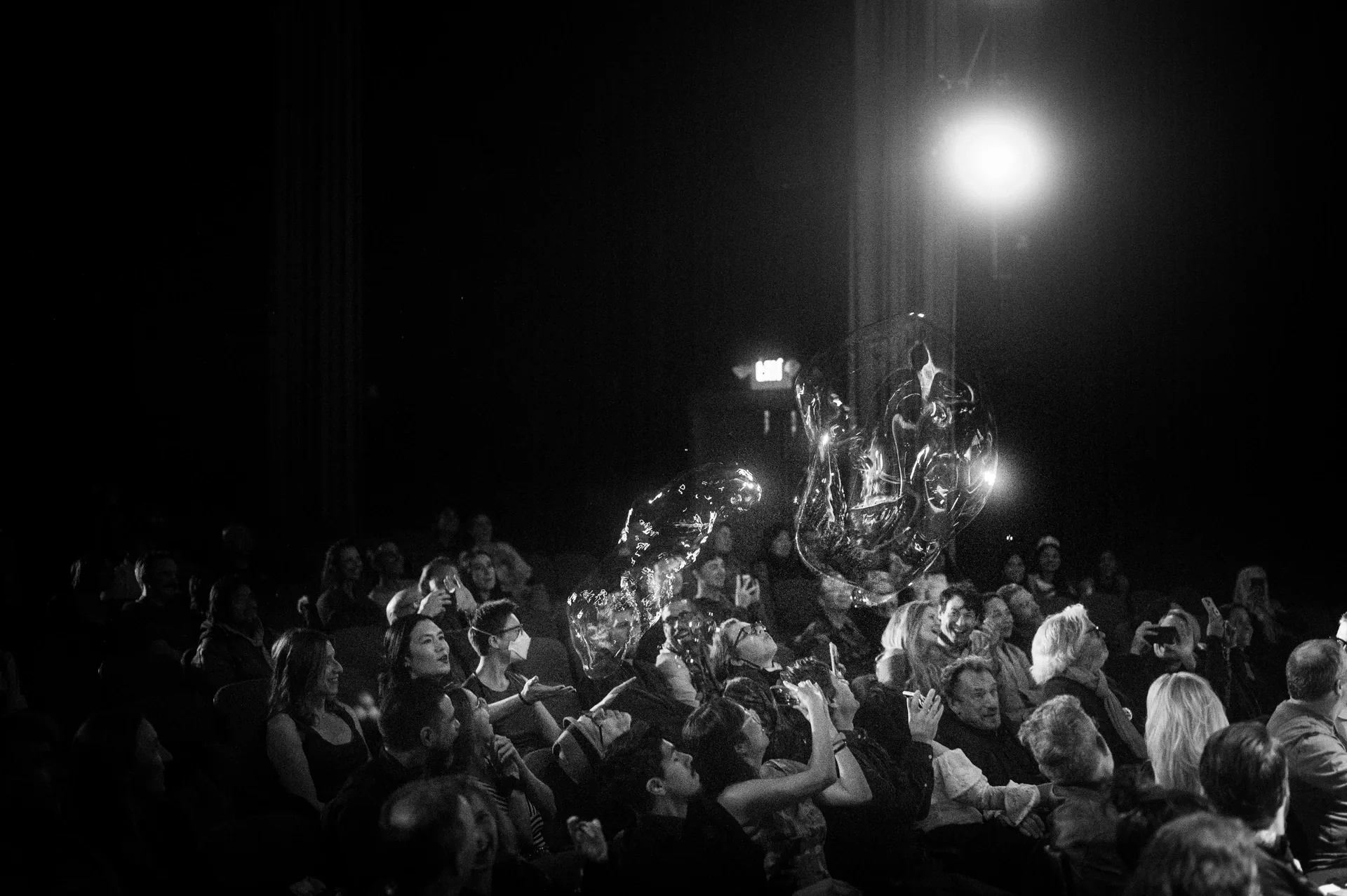 Costumed performer stands amid a seated audience under a bright backlight.