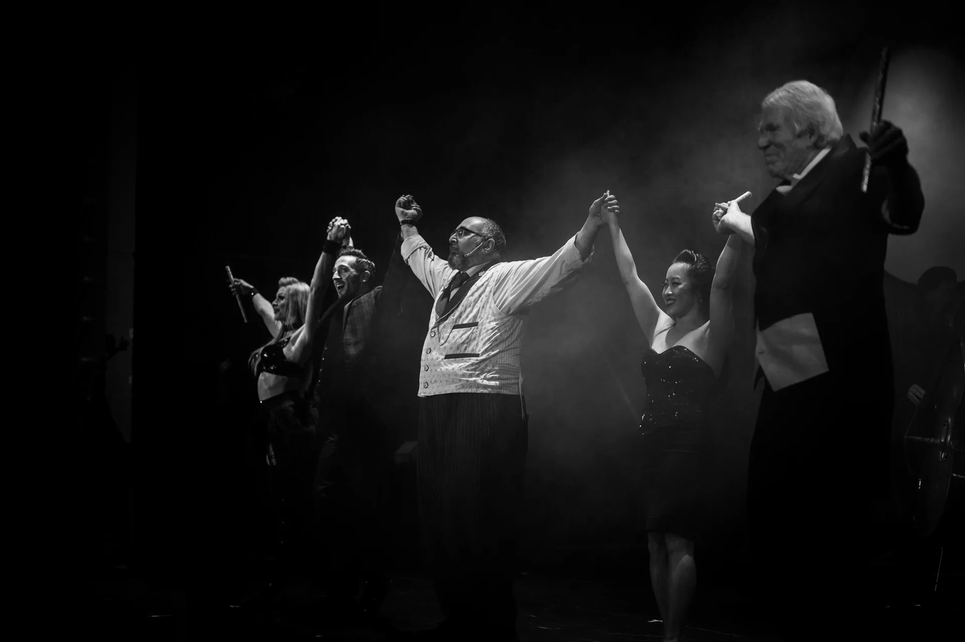 Singer raises both arms while dancers and a violinist perform behind.