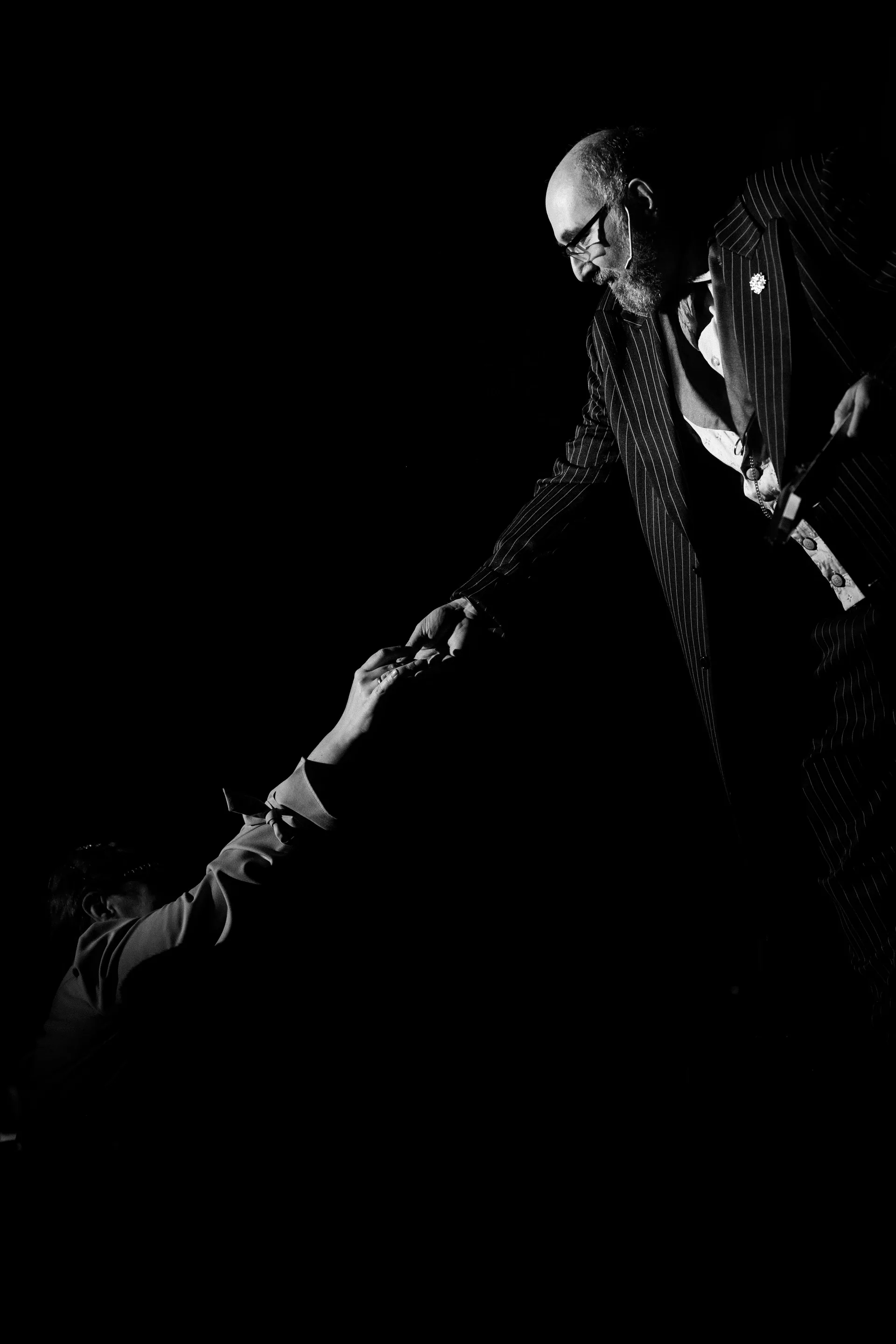 Suited performer holds the hand of a seated figure onstage.