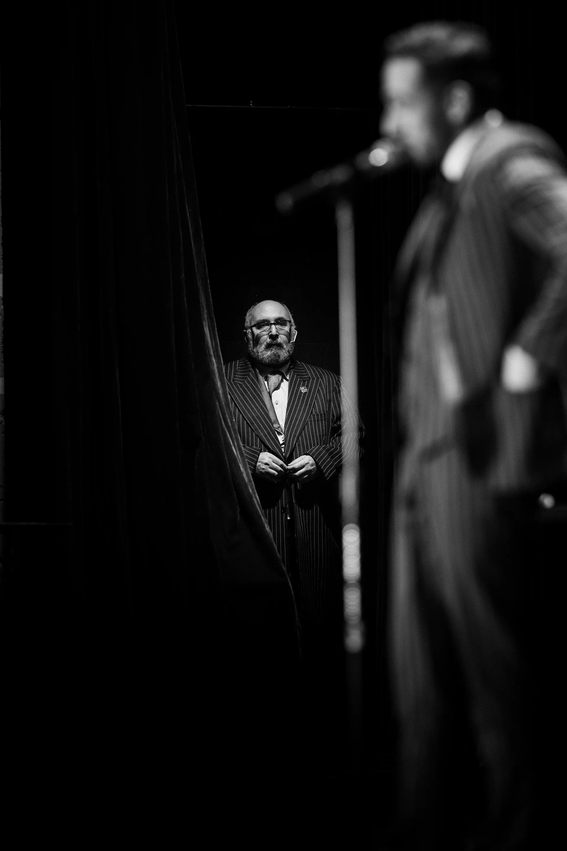 One performer stands at a microphone in the foreground while another watches behind.