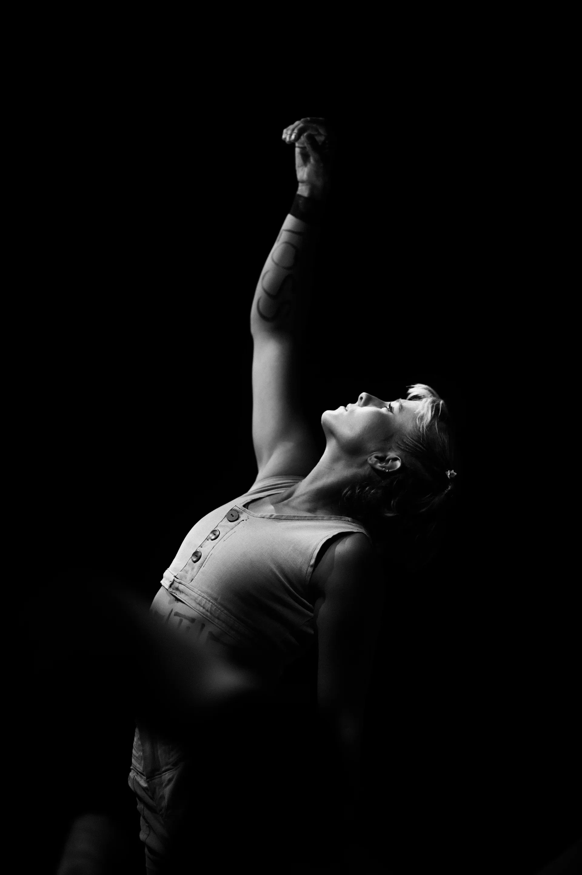 Performer leans backward with one fist raised in a narrow spotlight.