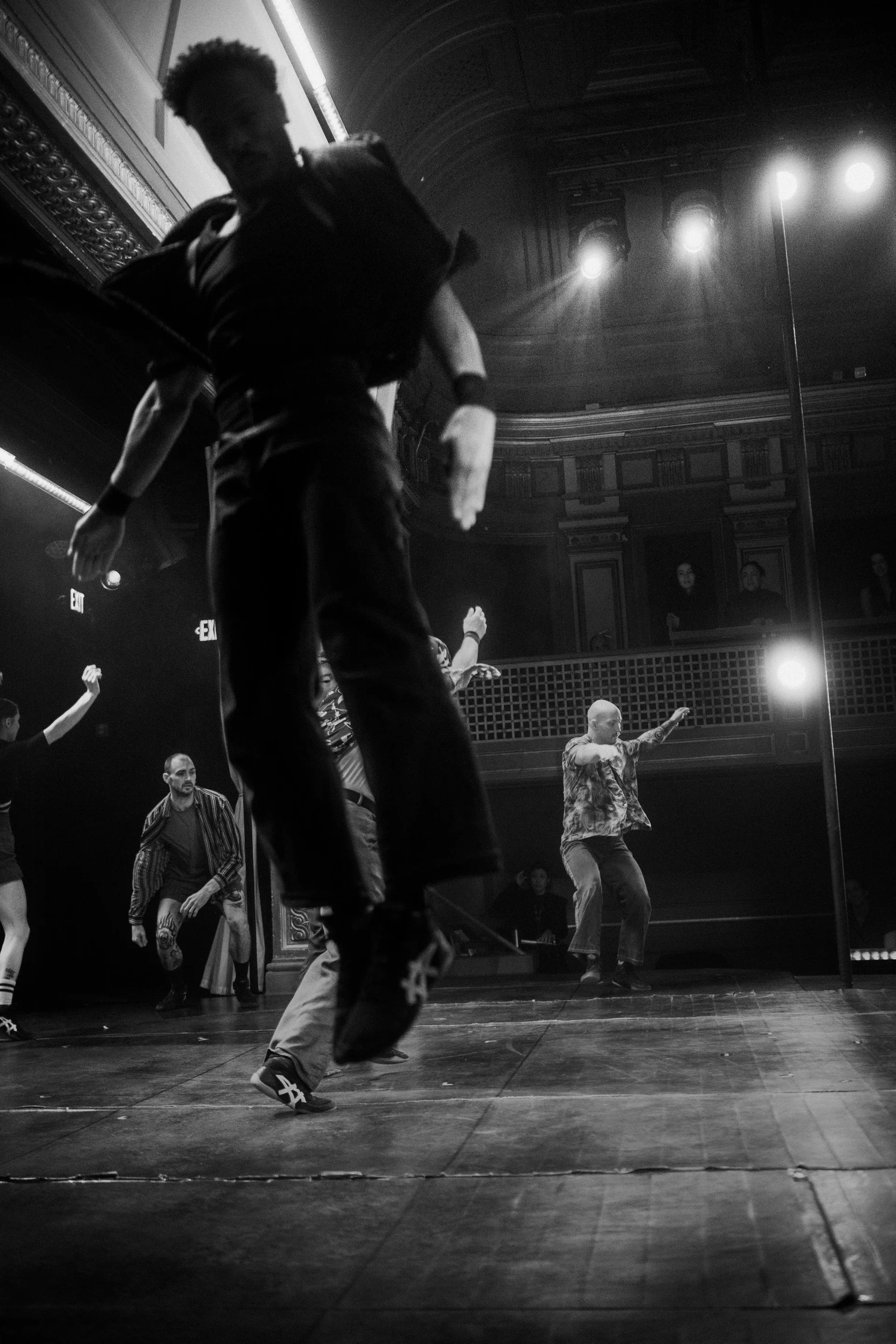 Performer walks across the foreground while an ensemble moves behind.