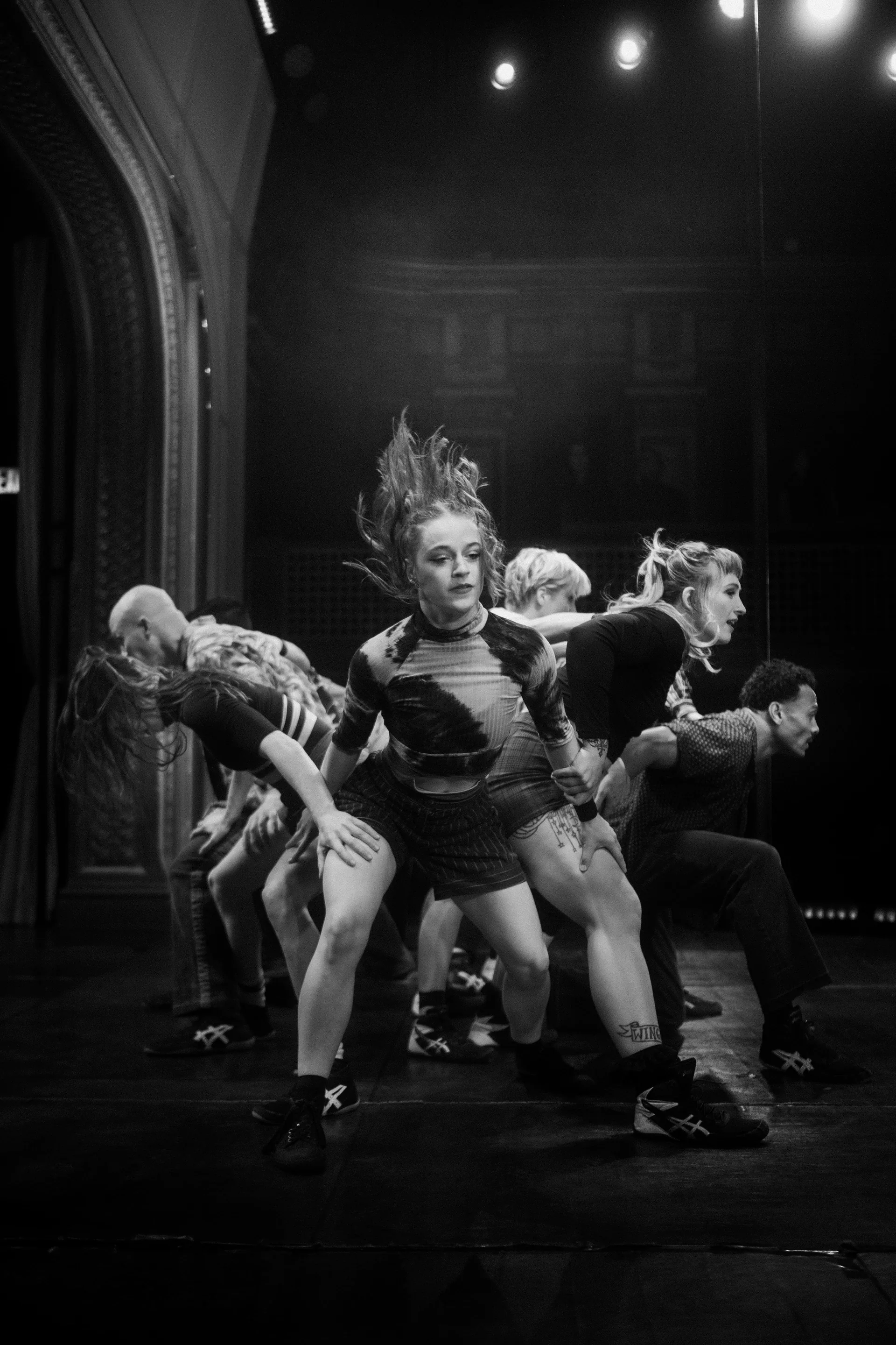 Performer crouches in the foreground while an ensemble bends behind.