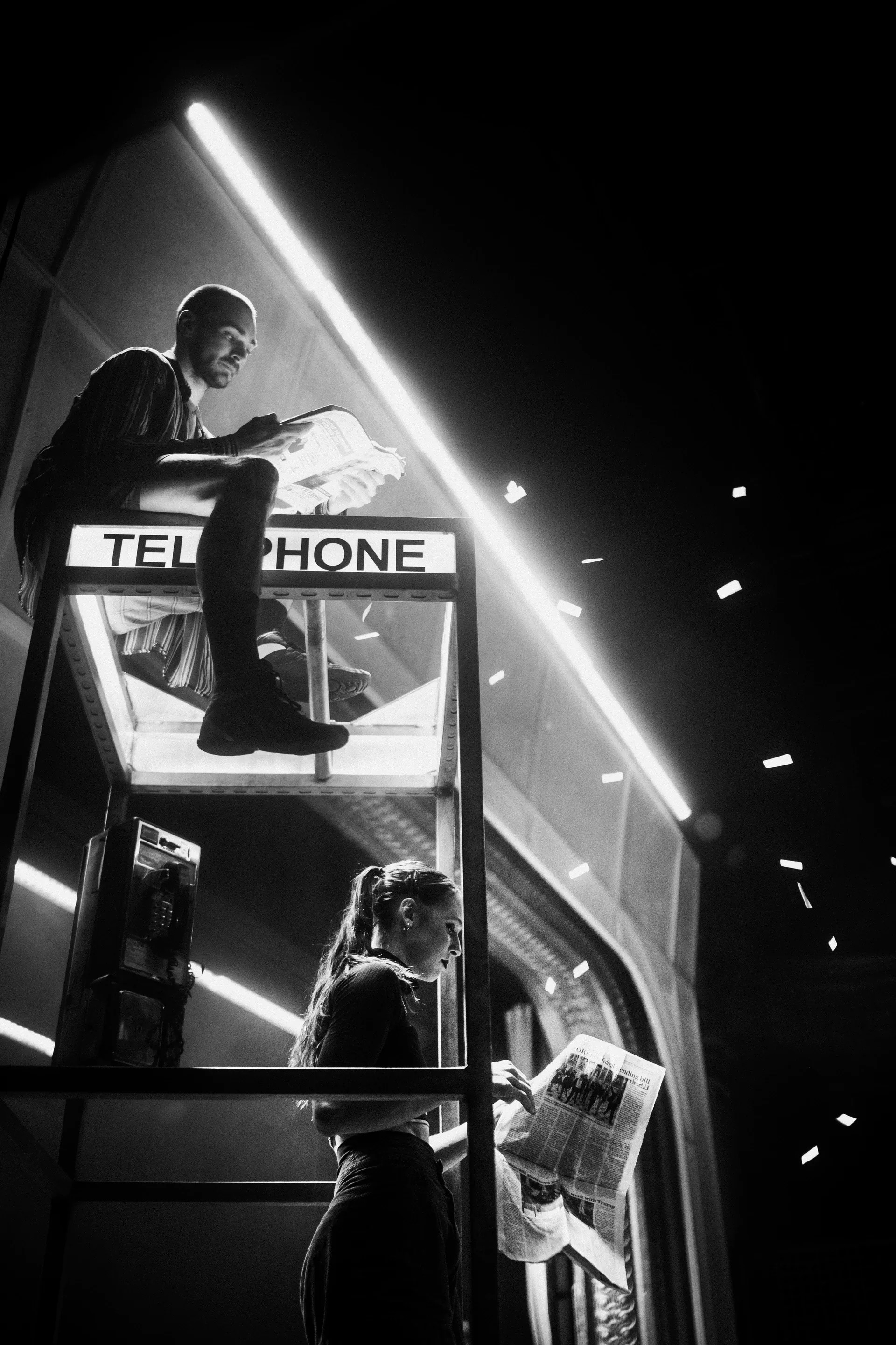 One performer sits atop an illuminated telephone booth while another climbs below.