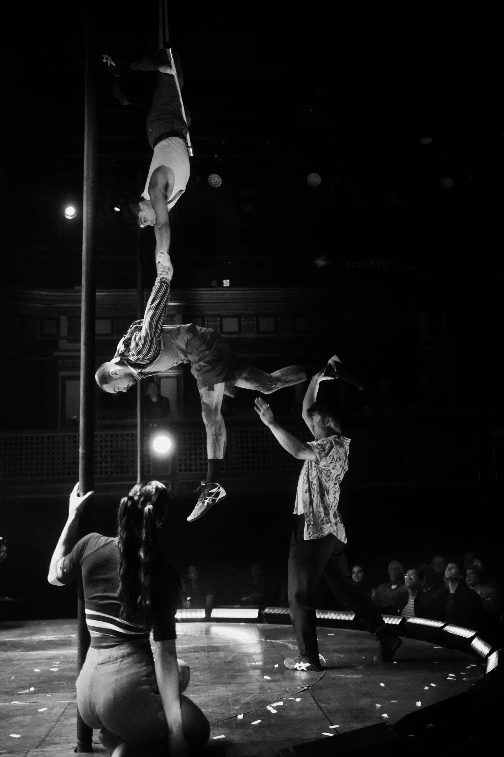 Acrobat hangs upside down above a partner and surrounding ensemble.