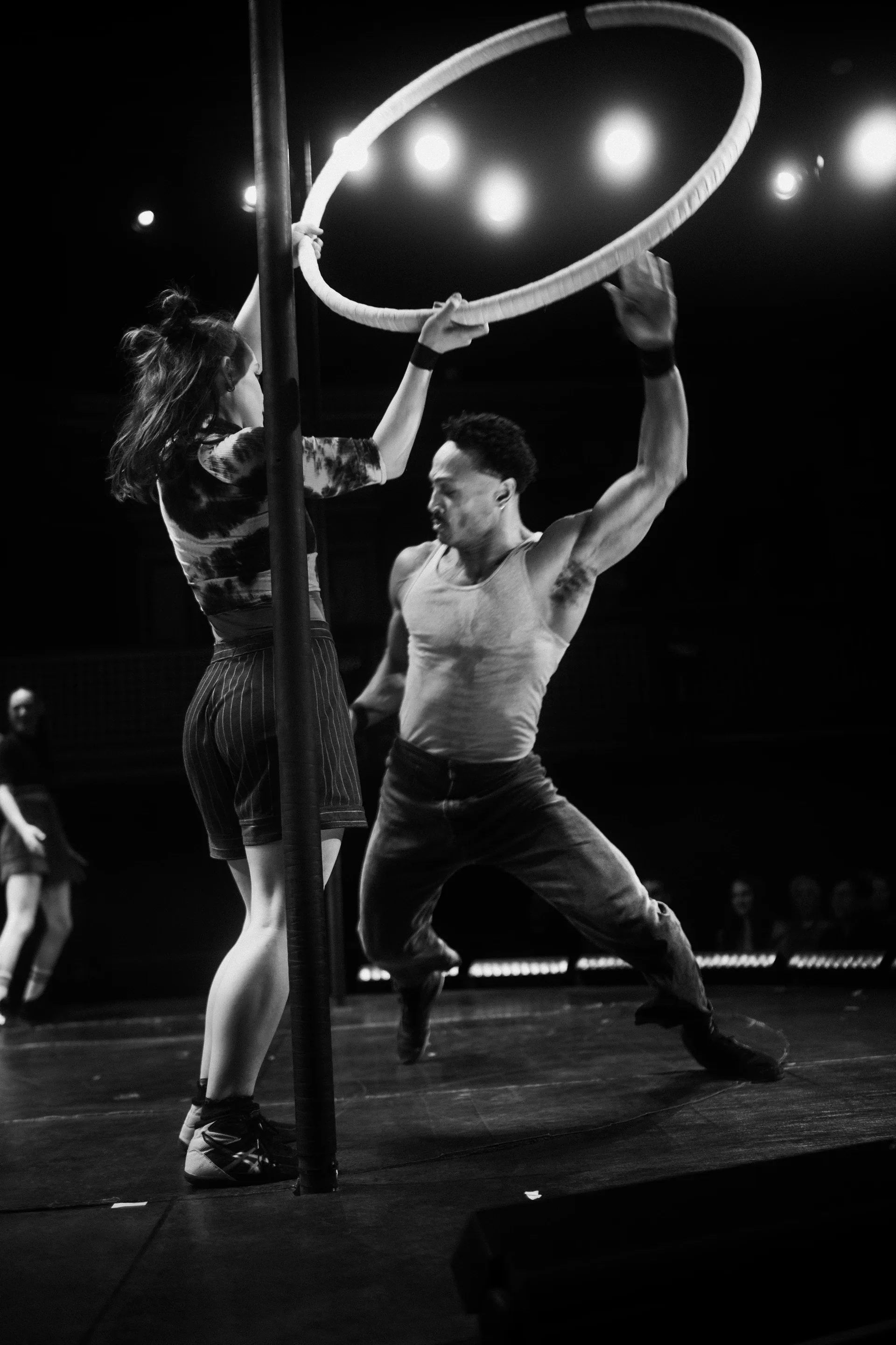 Two performers carry a large hoop upright while moving across the stage.