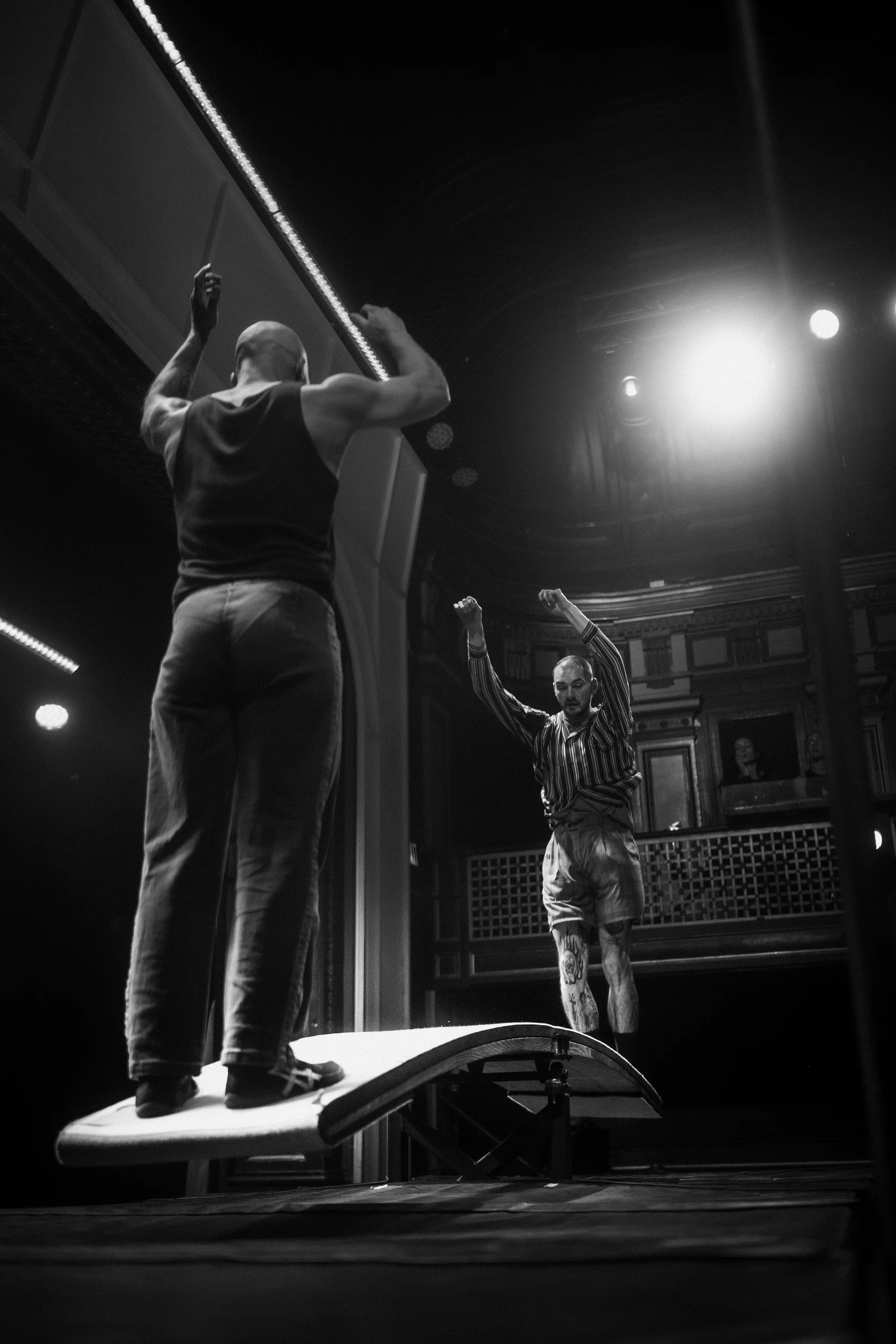 Two performers stand on a raised springboard while reaching upward.