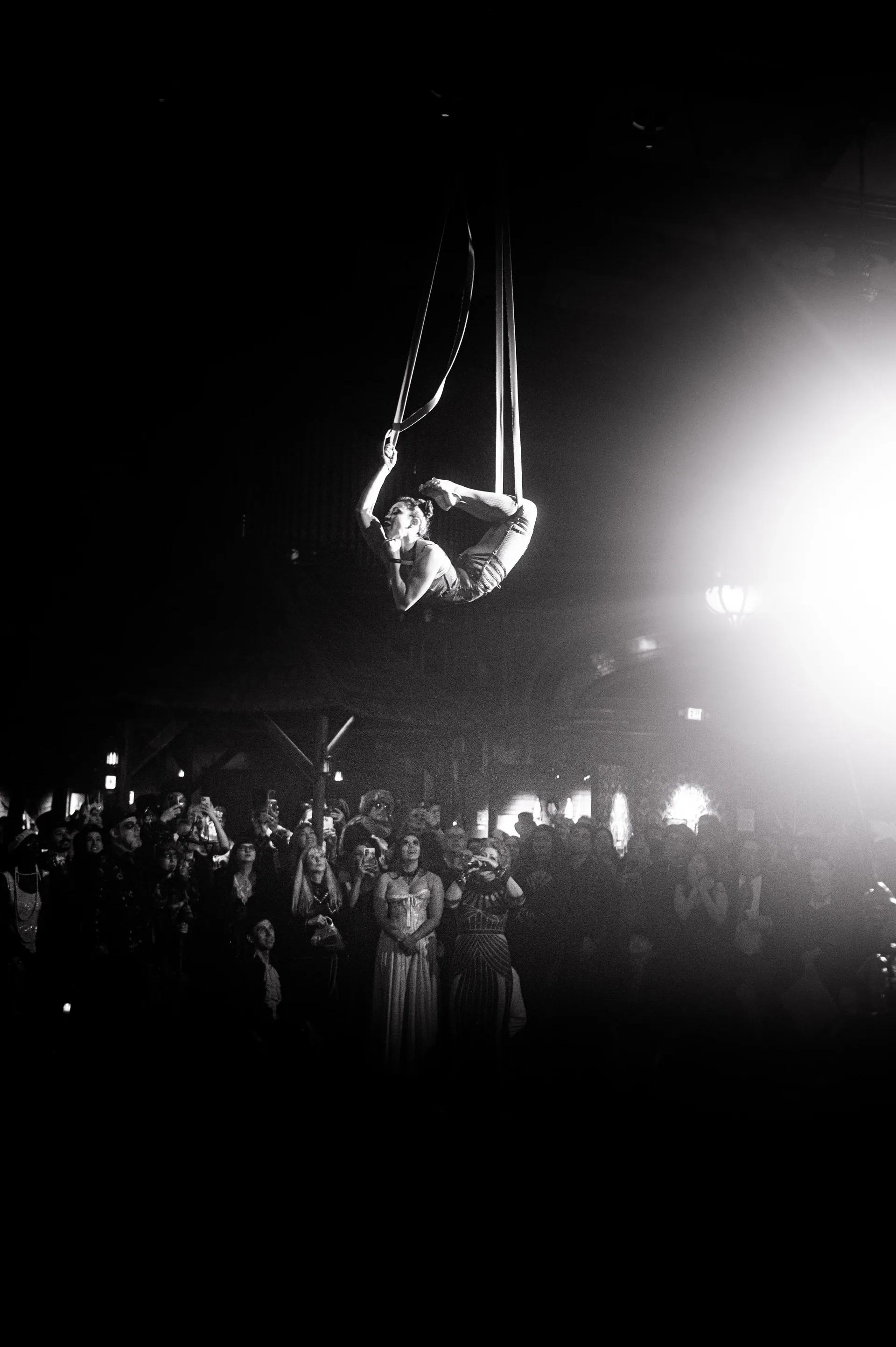 Aerialist hangs high above a dense crowd in a large hall.