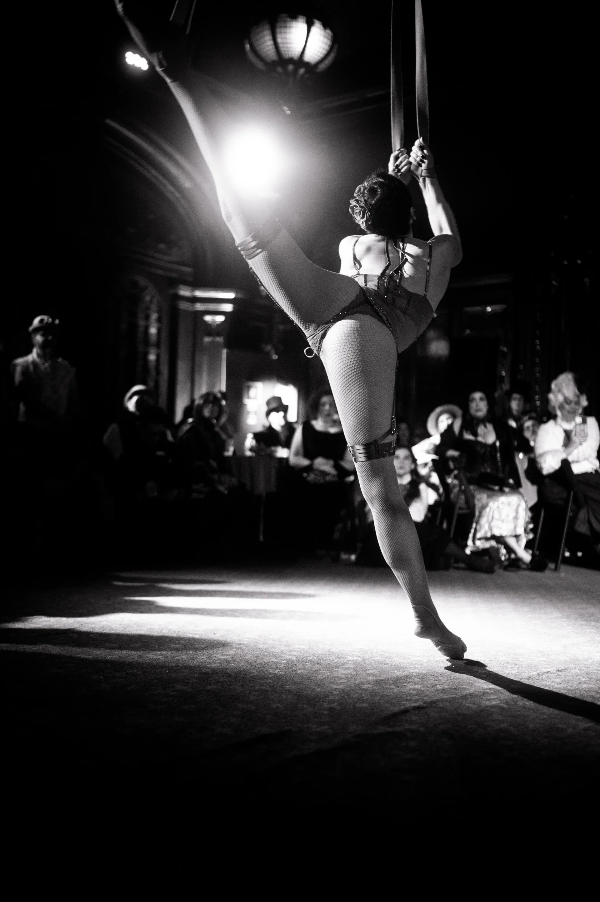 Aerialist poses on vertical straps above a seated audience.