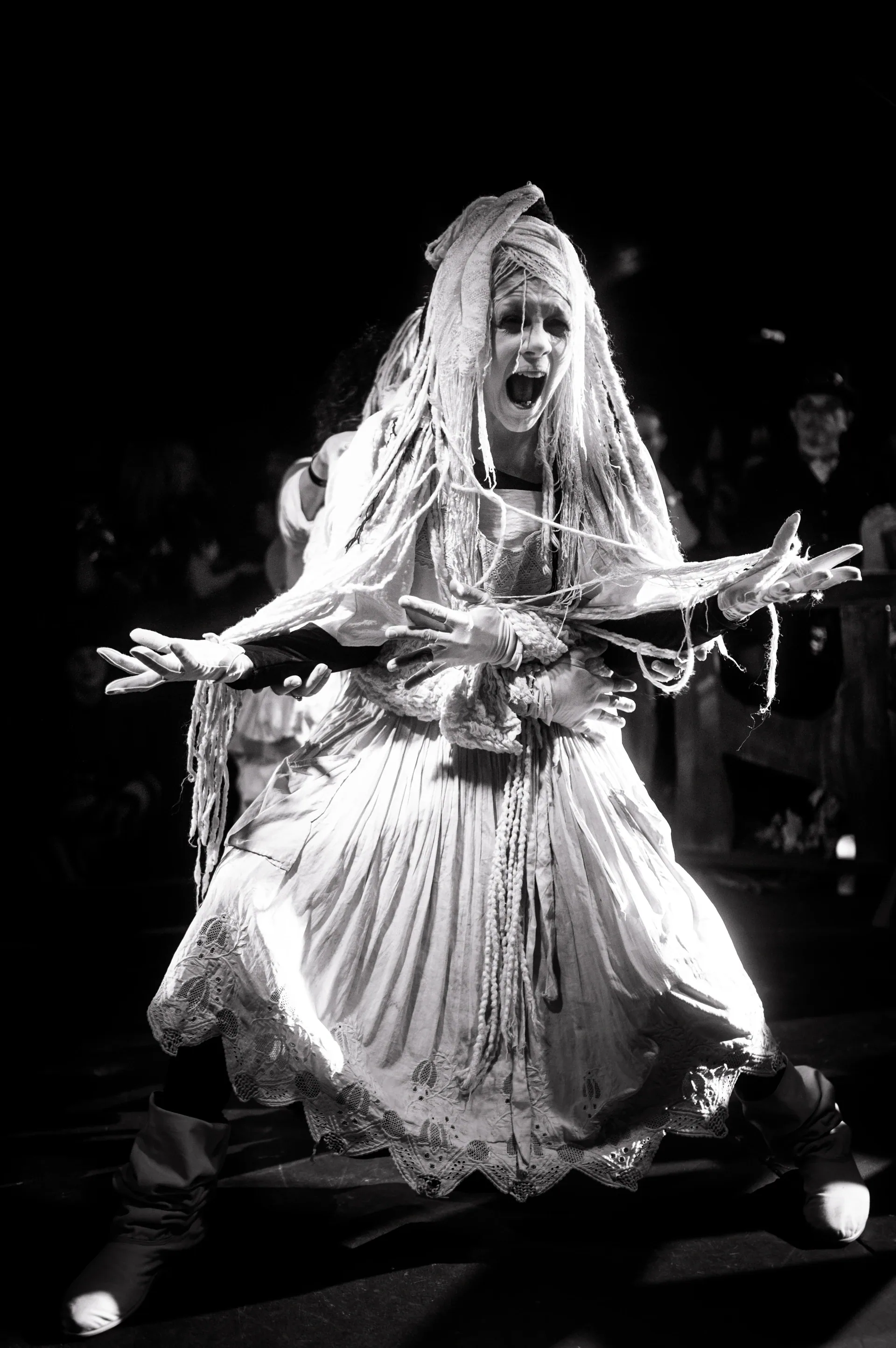 Performer in a layered, tattered costume crouches facing the camera.
