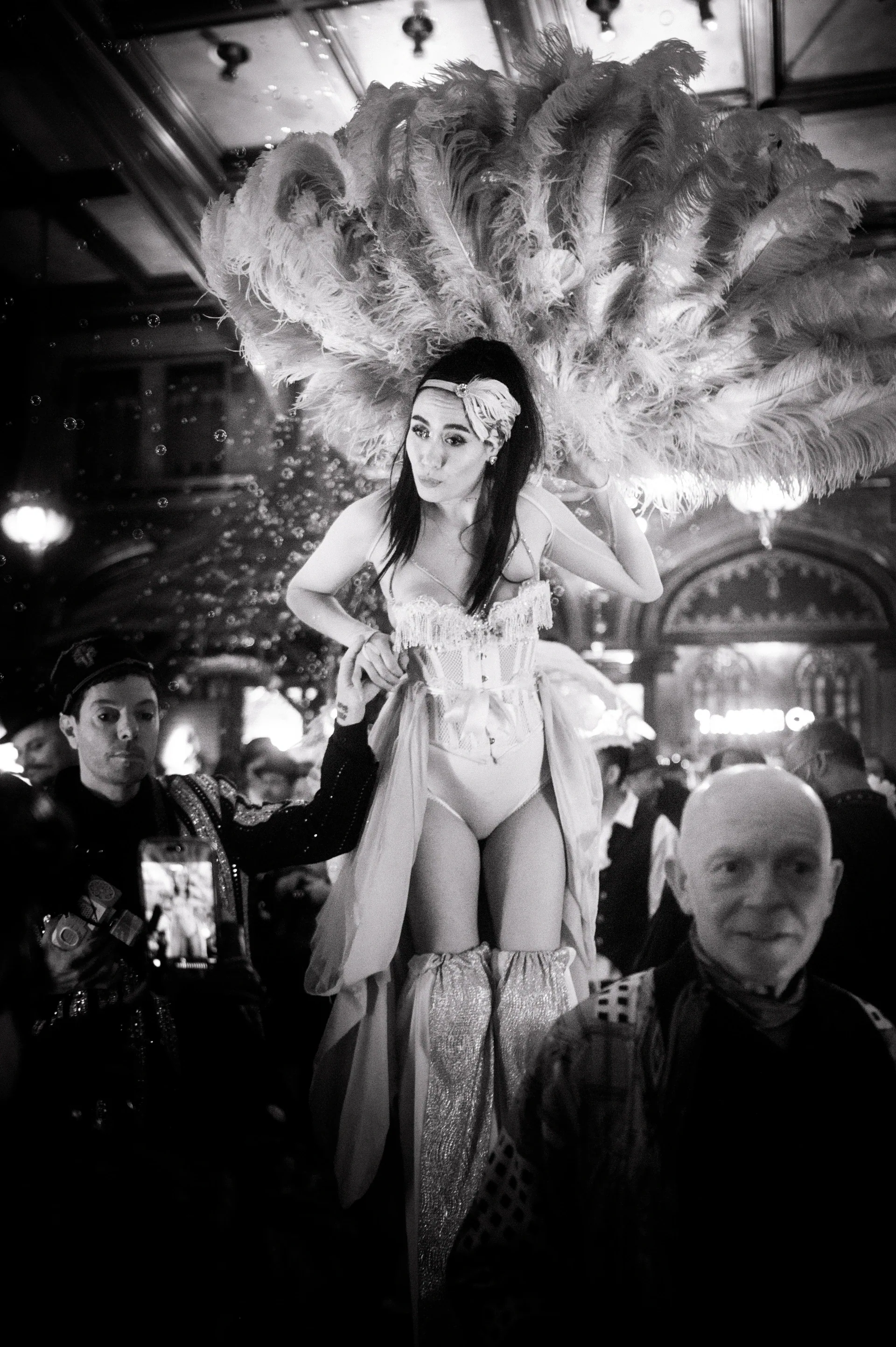 Performer wearing a large feathered headdress faces the camera amid a crowd.