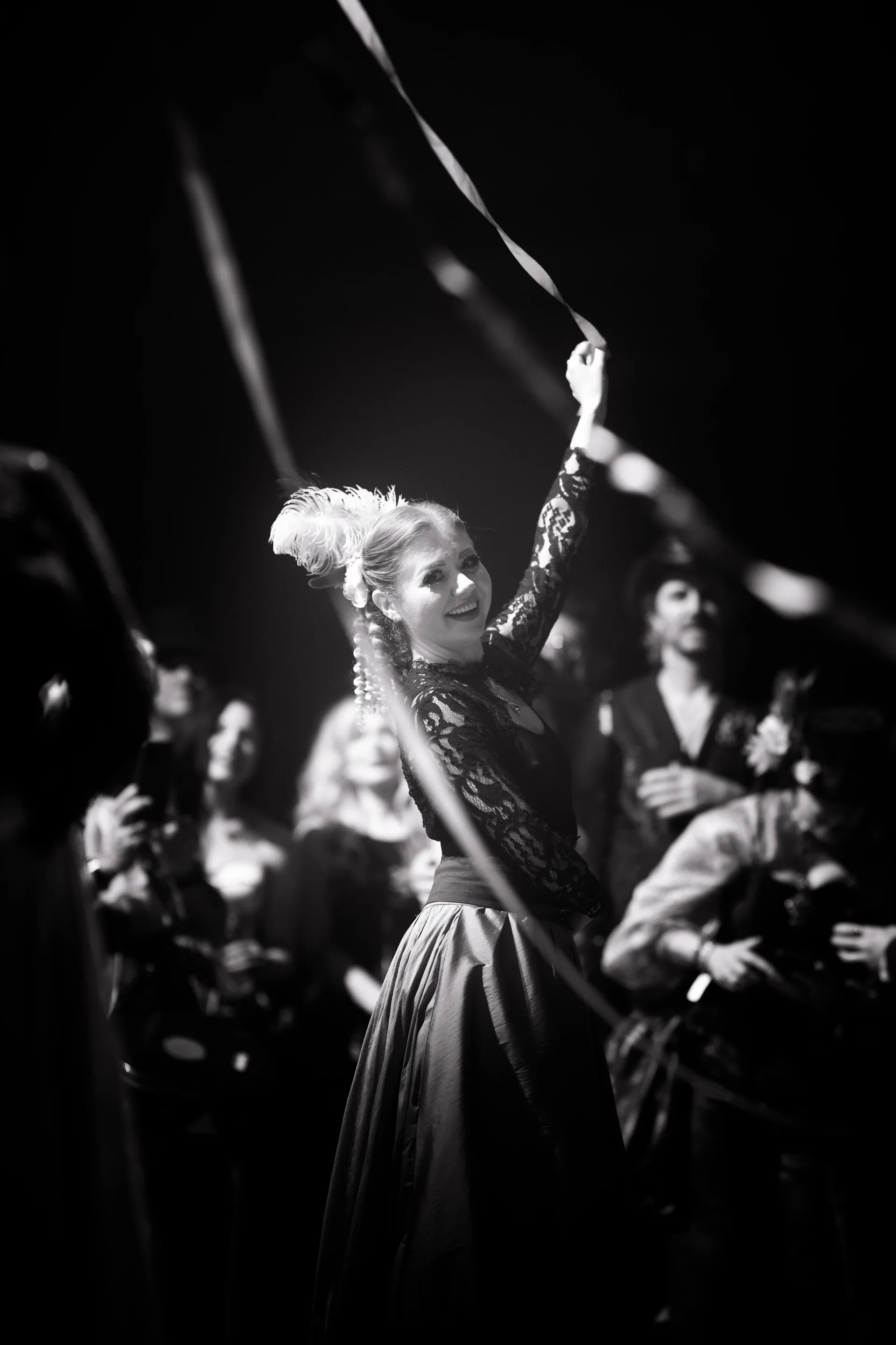 Performer in a long dark dress raises a tall ribboned pole among the crowd.