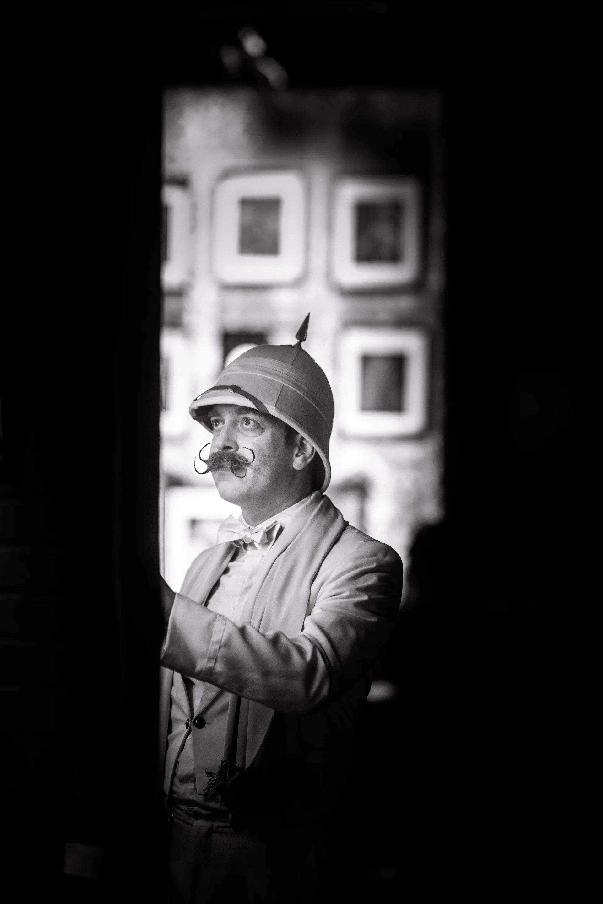 Person in a hat and suit stands in a doorway with hands together.