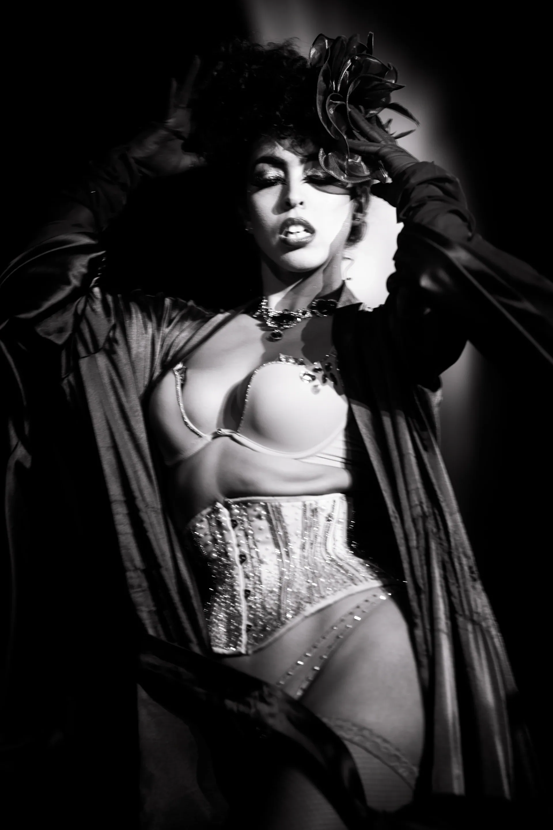 Performer in a corset and feathered headpiece poses beneath a spotlight.