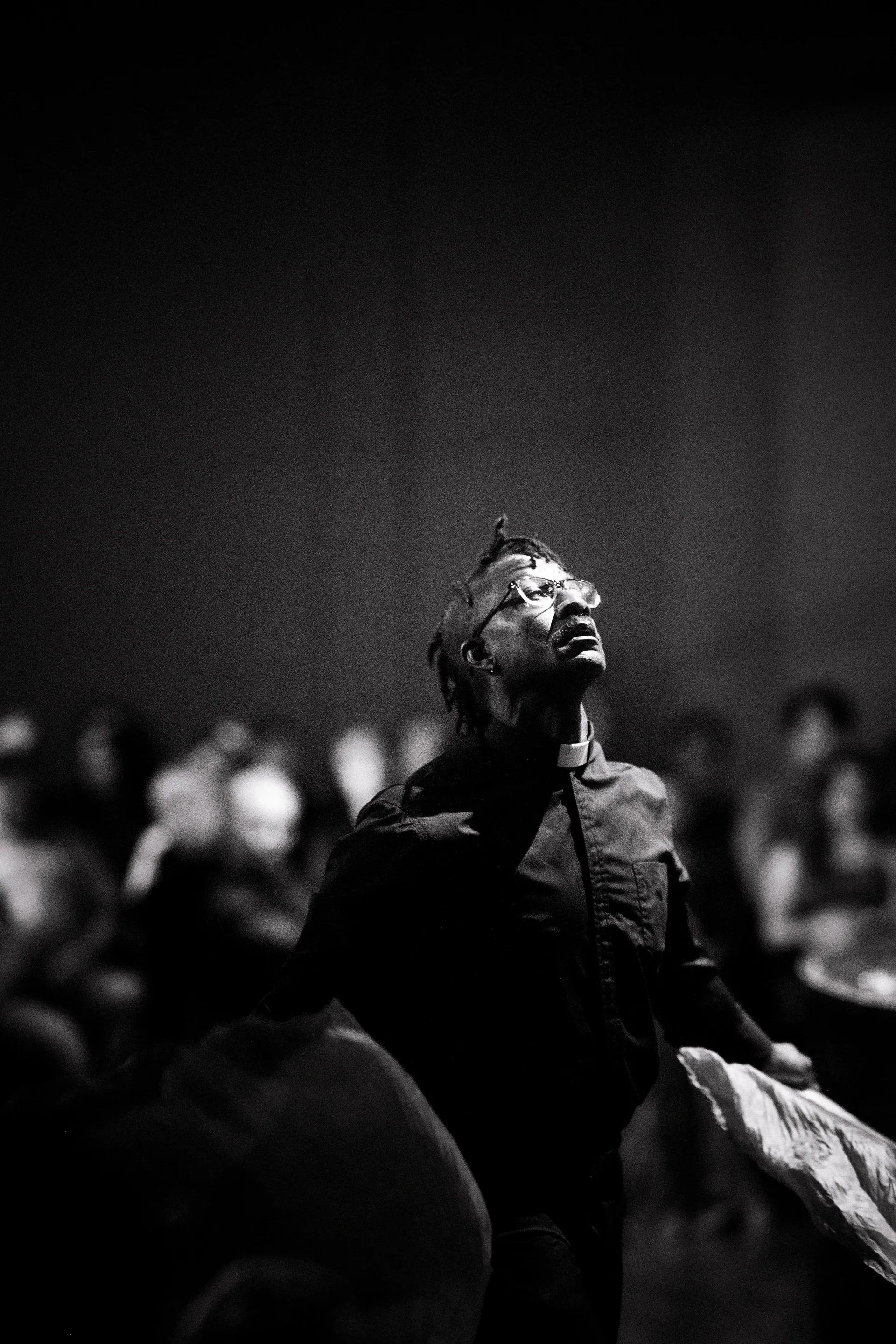 Suited performer sits among an audience and looks upward.