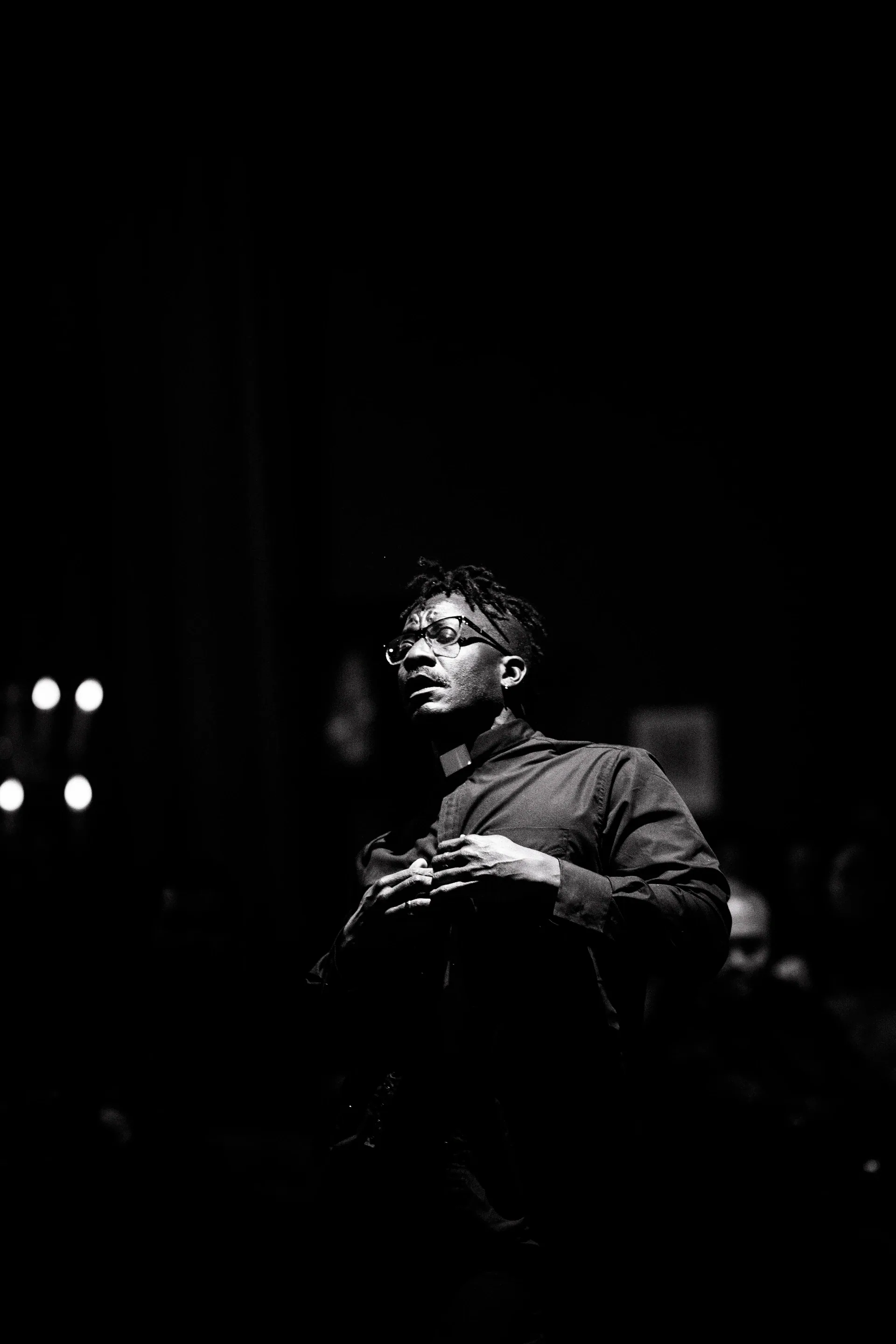 Suited performer stands in the dark with both hands held near the chest.