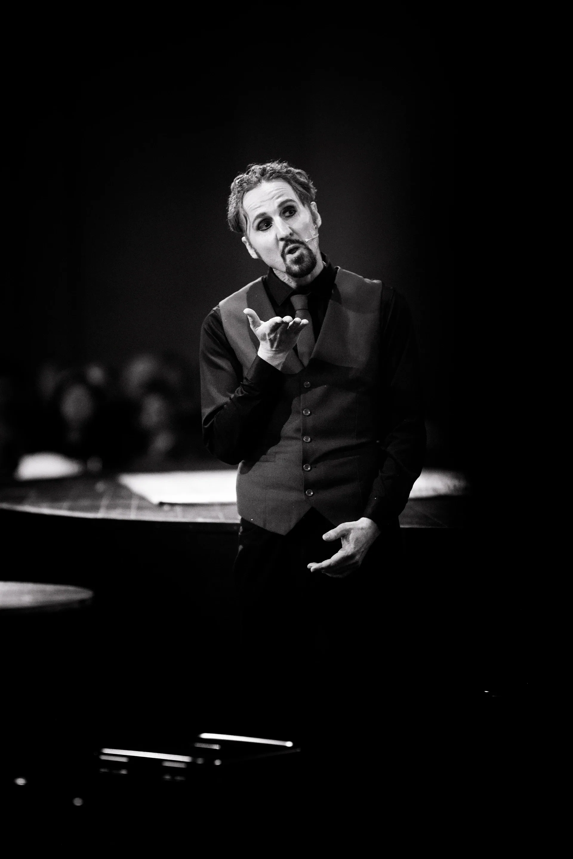 Suited performer leans against a counter and faces the audience.