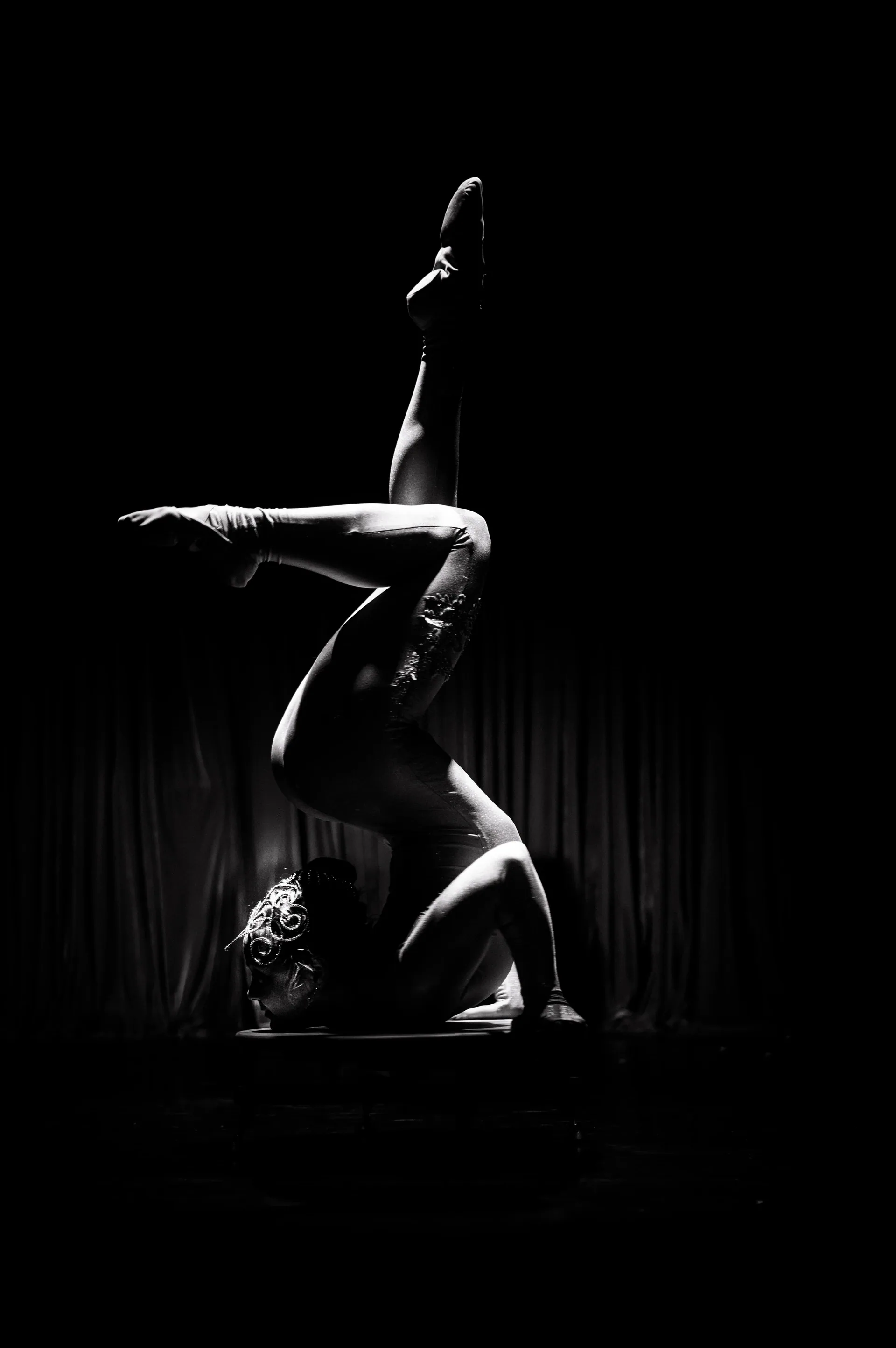 Hand-balancing performer holds the legs bent overhead against a dark backdrop.