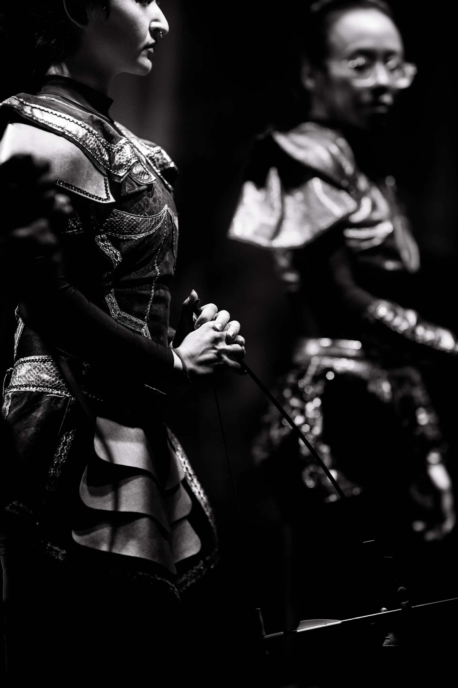 Performer in an armor-like costume stands in profile with a musician behind.