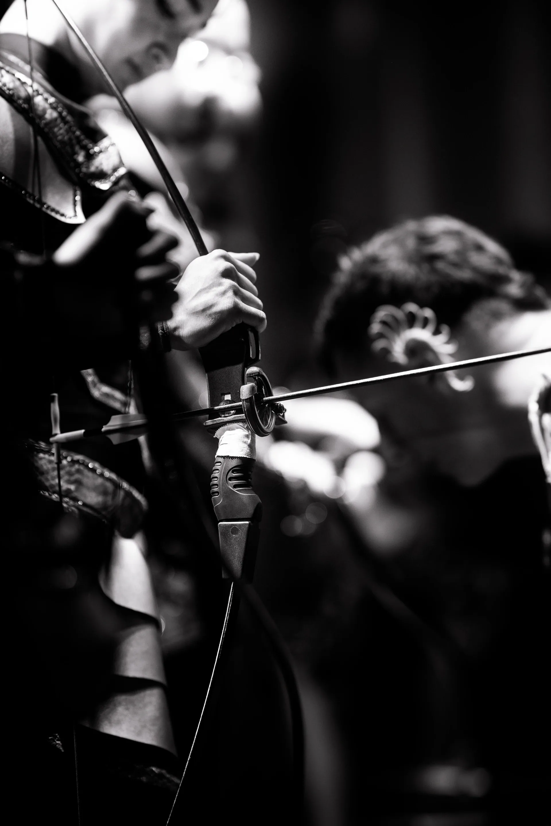 Close view of a performer gripping a compound bow with another person blurred behind.